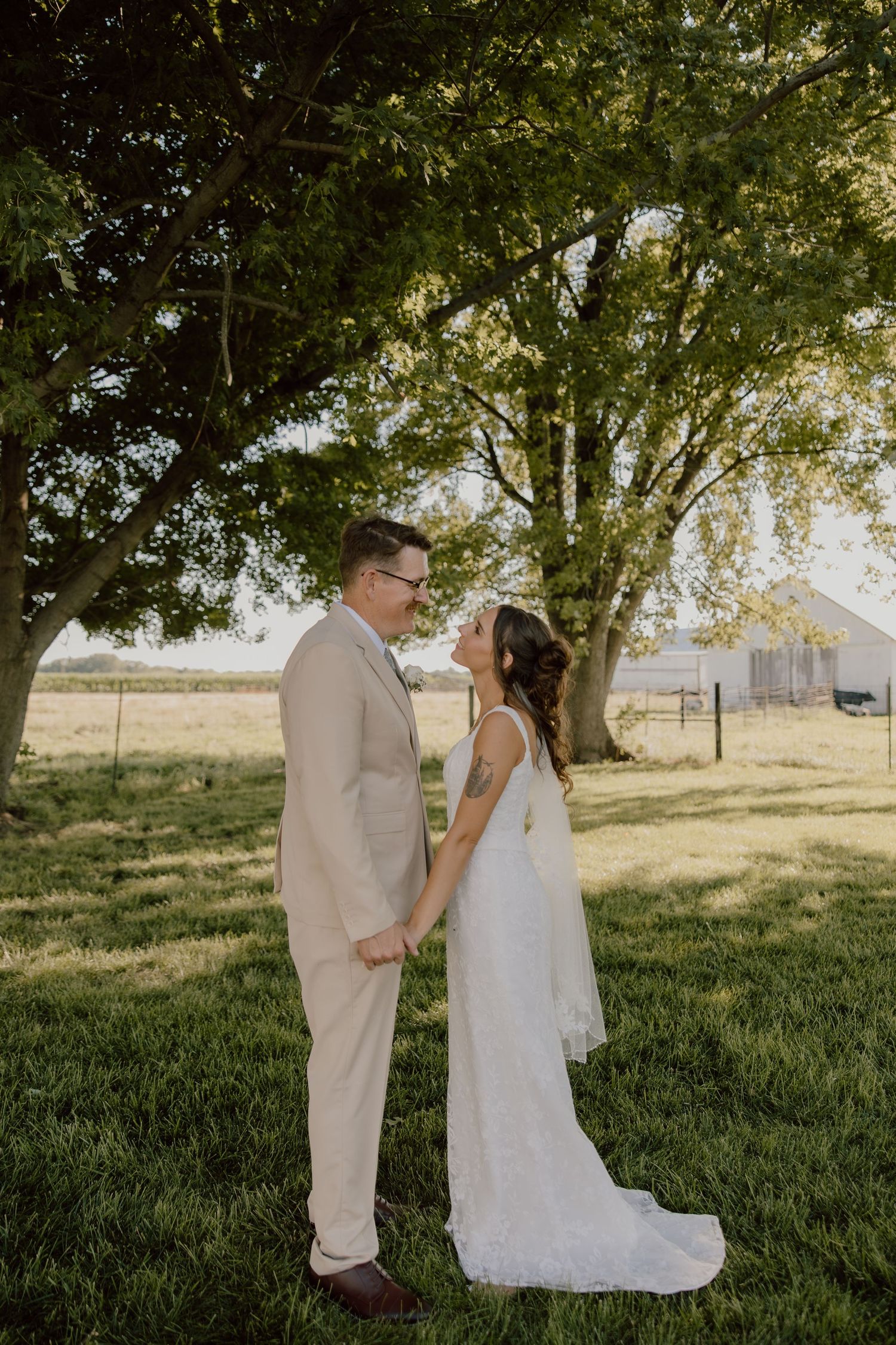 A bride and groom hold hands and gaze at each other under a large tree on a green lawn at a rustic farm wedding.