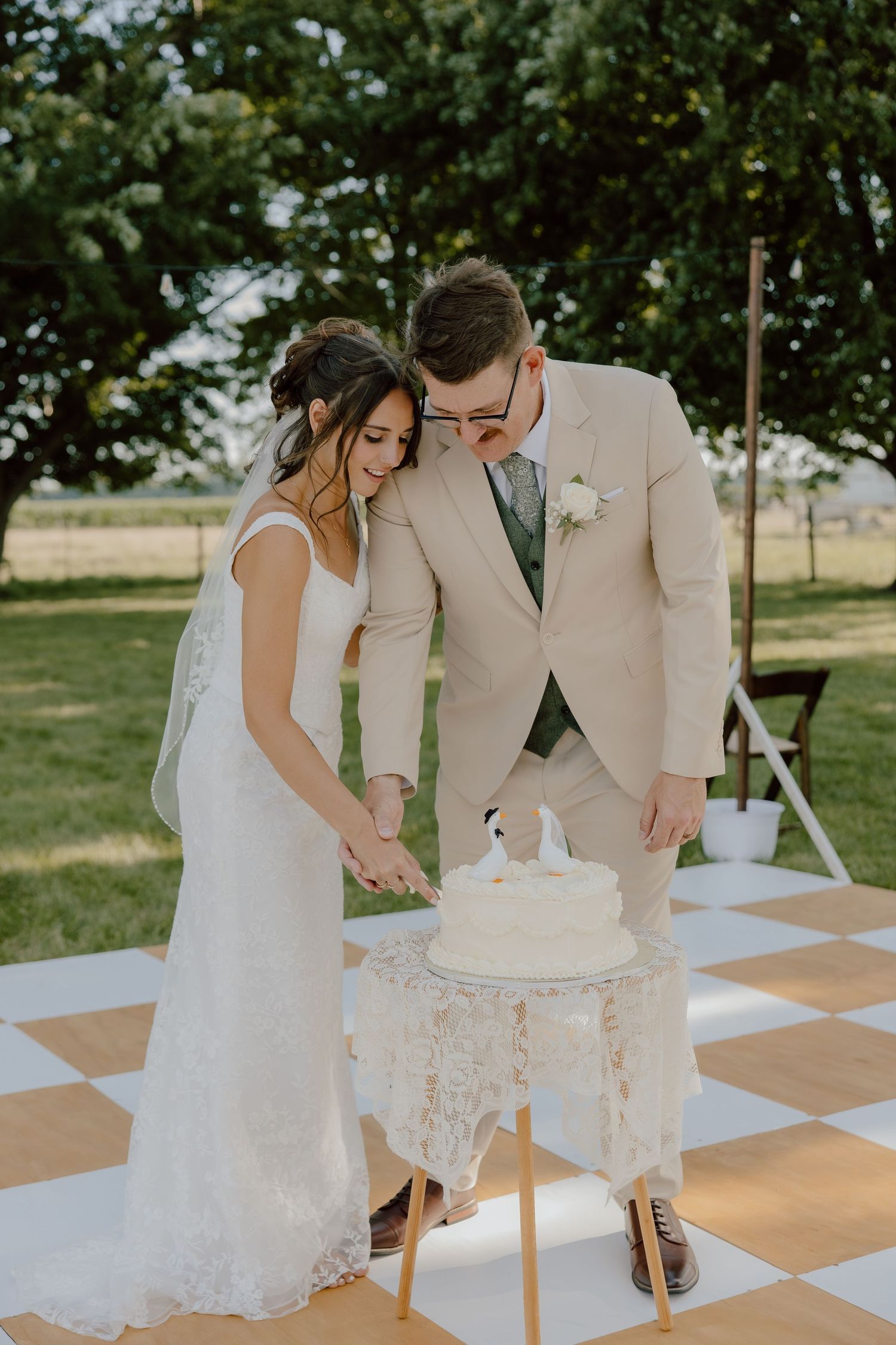 Bride and groom cutting white wedding cake outdoors on checkered dance floor surrounded by lush green trees.