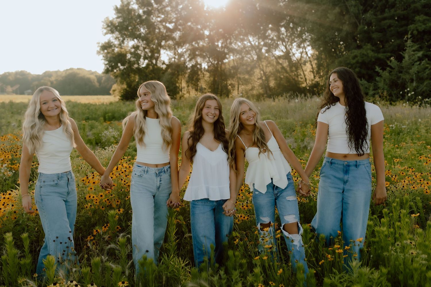 Five young women holding hands in a sunlit wildflower field, wearing white tops and jeans, smiling outdoors.