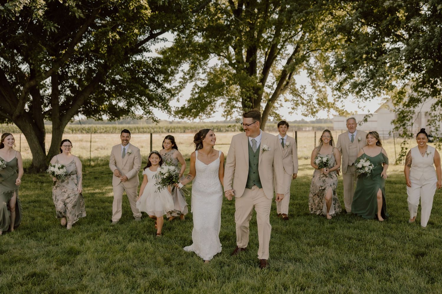 Wedding party walks outdoors on green grass under trees, bride in white gown with groom in tan suit, bridesmaids in sage green.