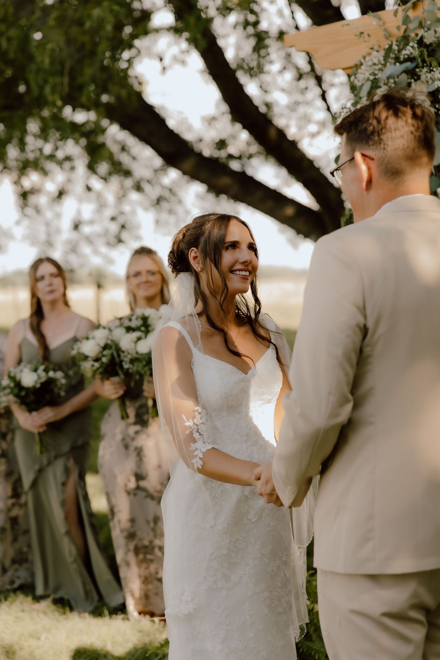 Smiling bride in lace wedding dress holds grooms hands at outdoor ceremony with bridesmaids in sage green.