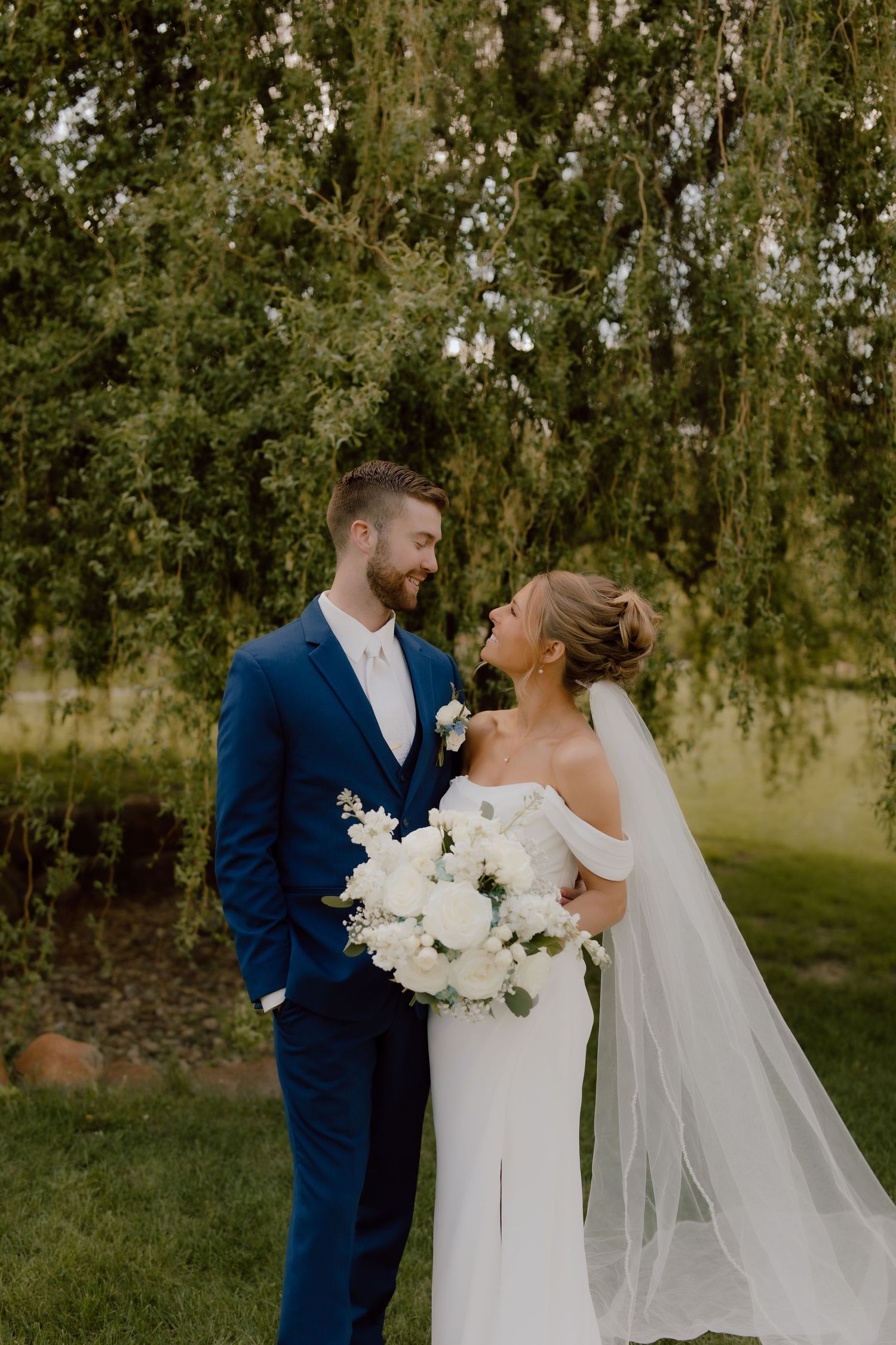 Bride and groom share a romantic moment under a weeping willow tree, bride holding white floral bouquet with veil flowing.