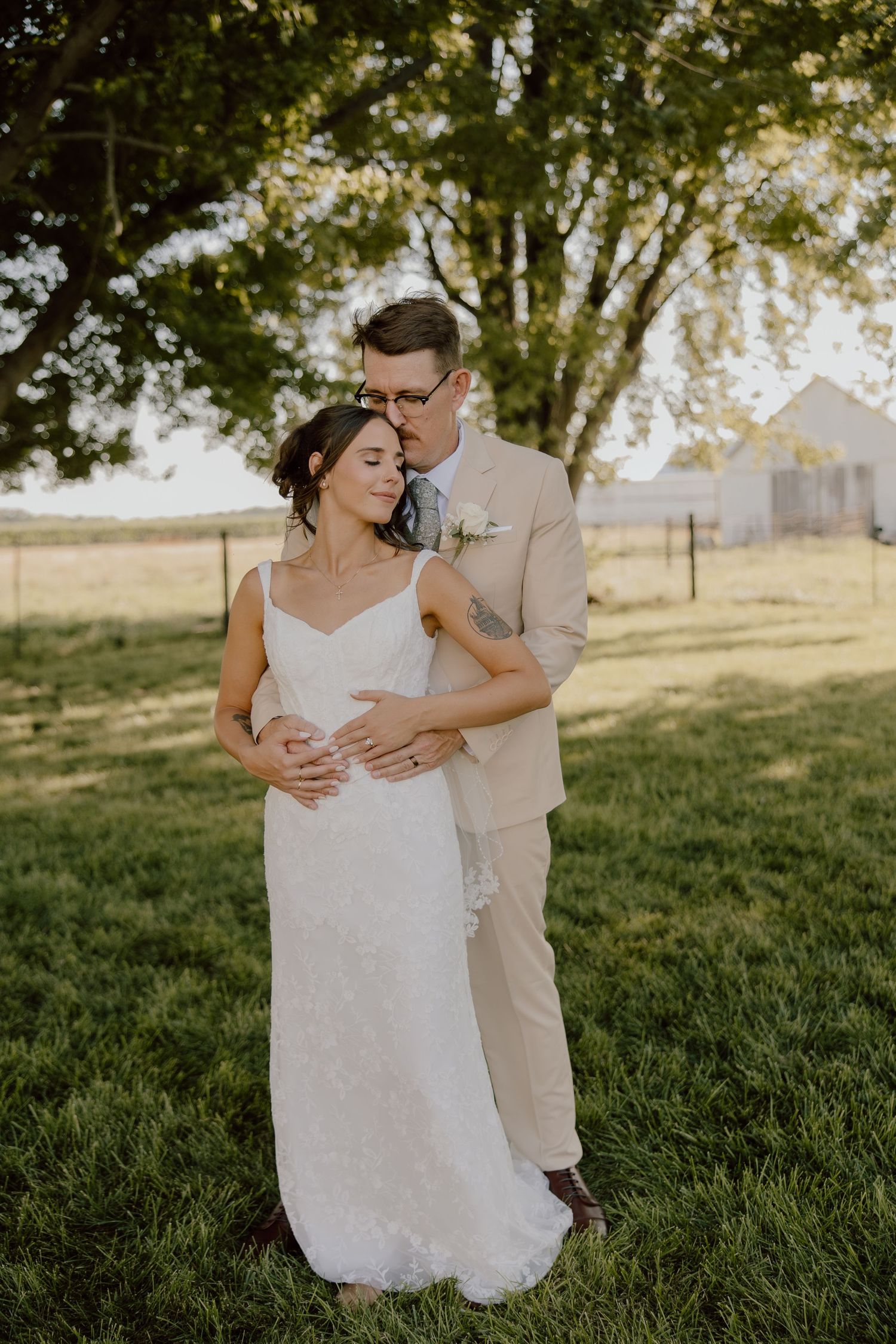 Bride and groom embrace in a sunlit green field, she in lace gown, he in tan suit with boutonniere.