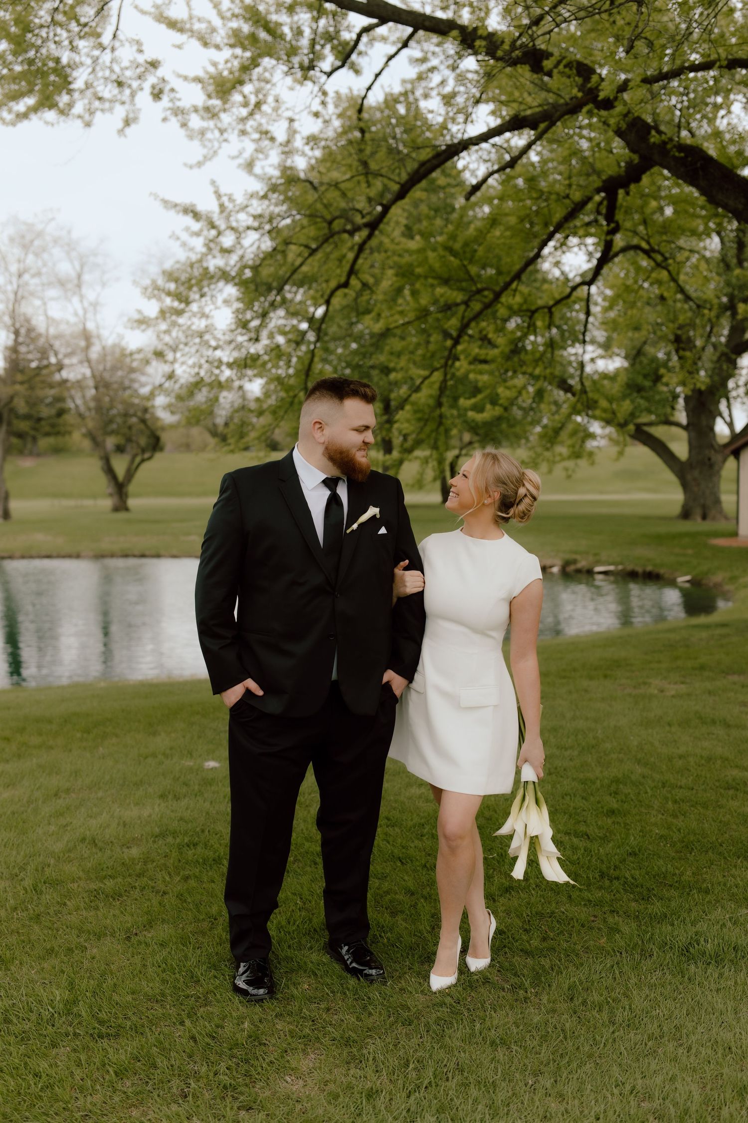 Couple in wedding attire standing by a pond, groom in black suit, bride in short white dress holding bouquet.