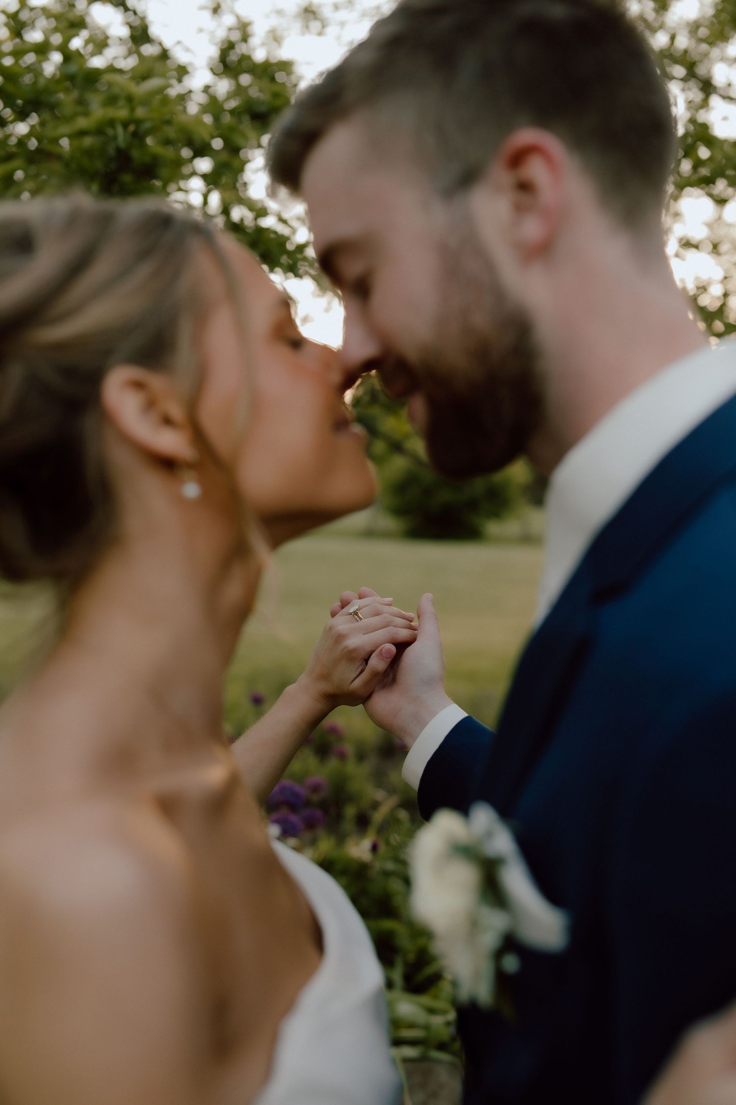 Bride and groom sharing a romantic kiss while holding hands outdoors, with soft bokeh greenery background.