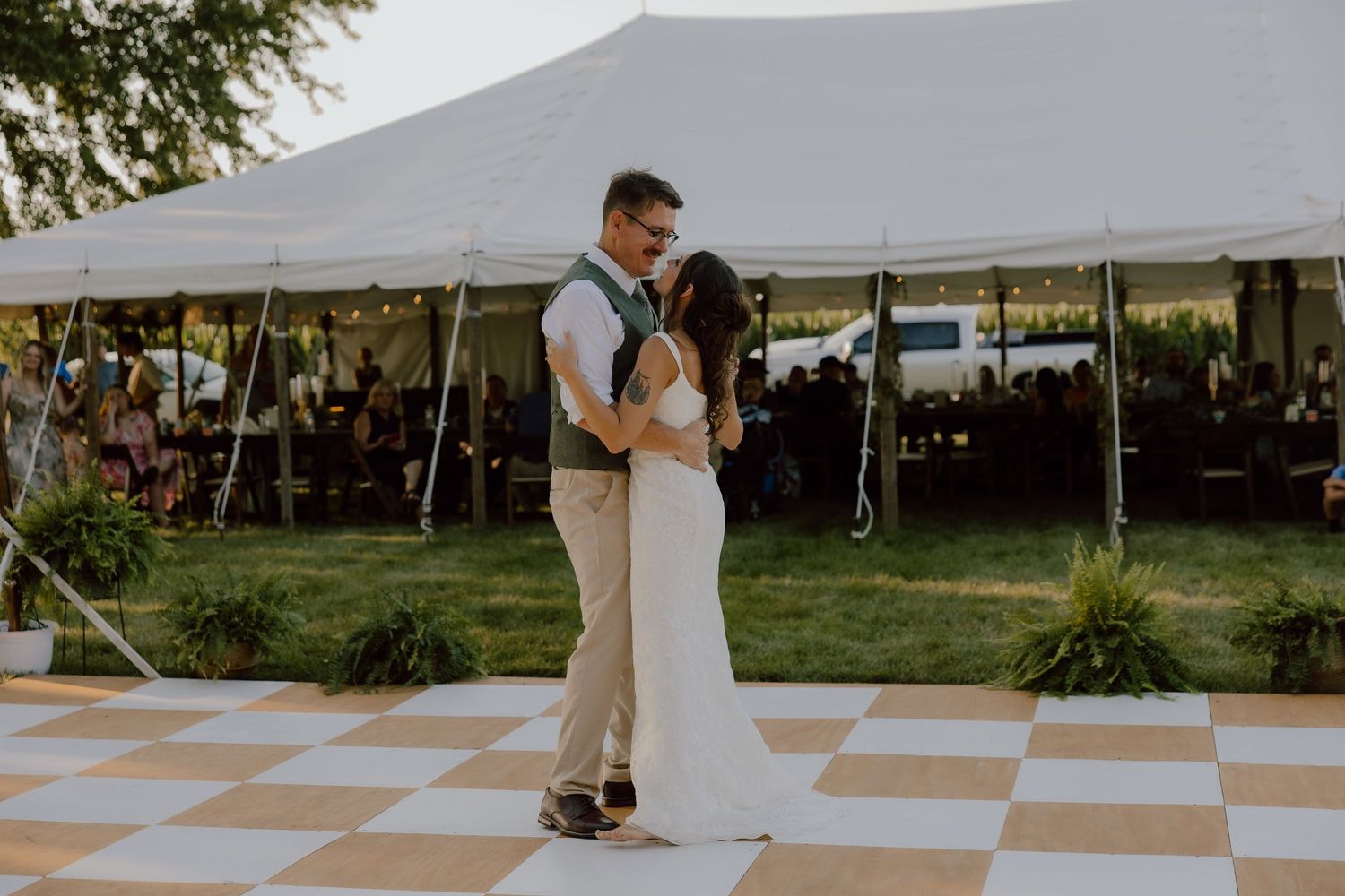 Bride and groom share first dance on checkered floor outside white tent at outdoor wedding reception.
