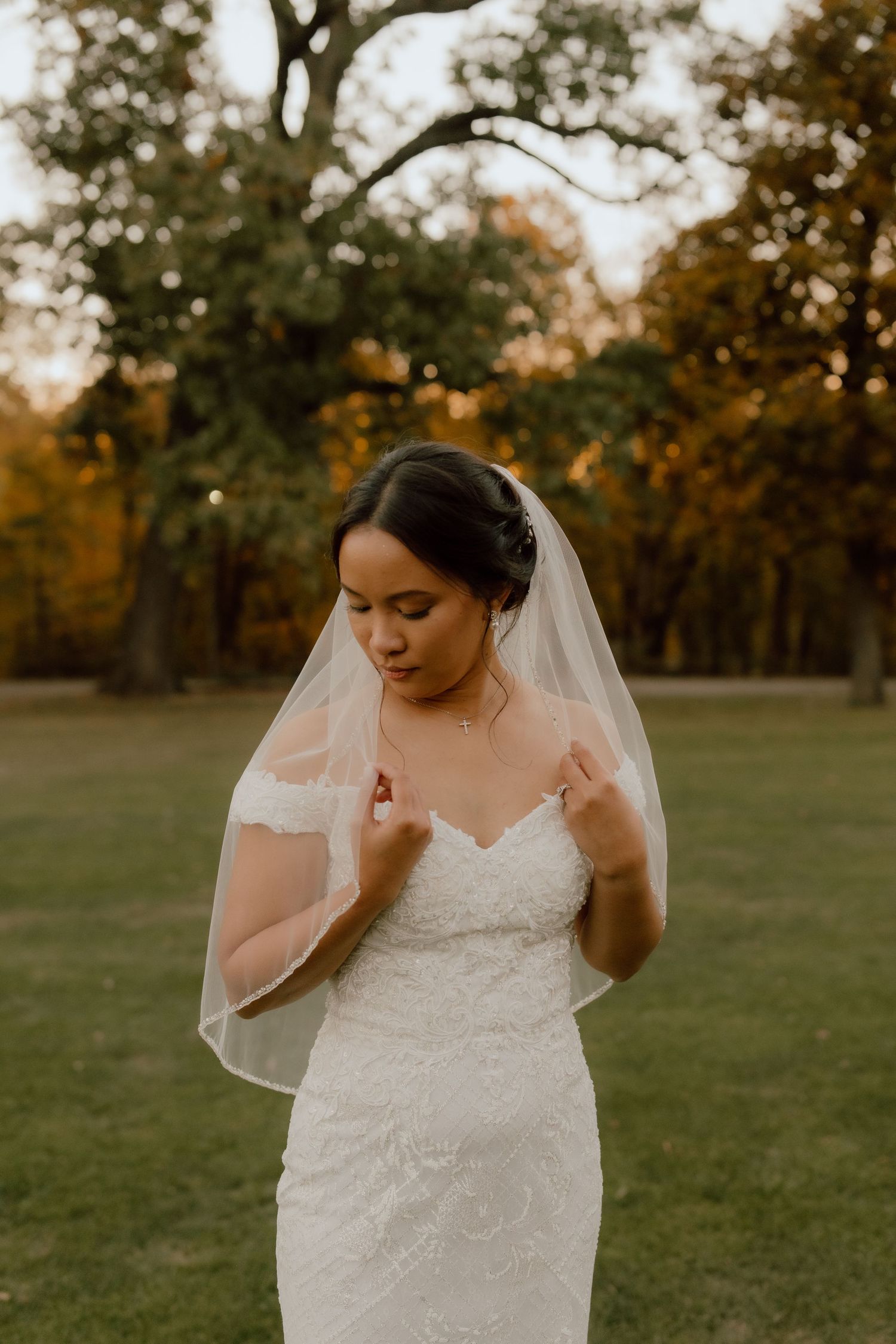 Bride in lace off-shoulder gown and veil poses outdoors during golden hour fall wedding portrait.