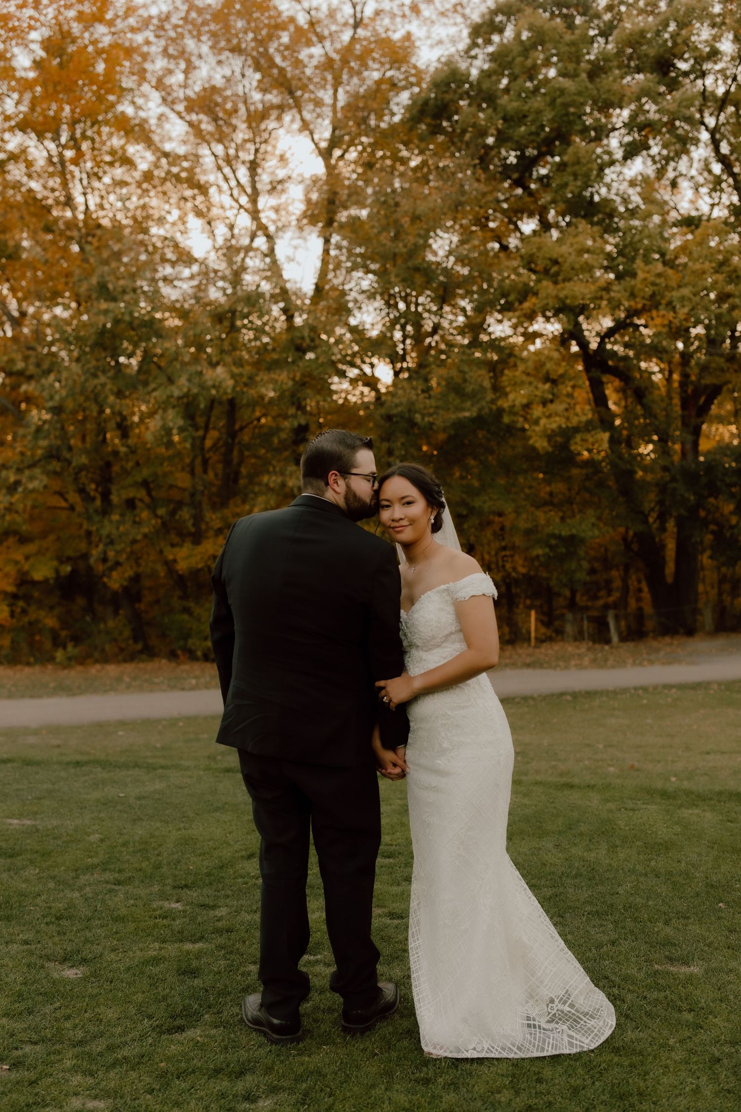 Bride in off-shoulder lace gown and groom in black suit embrace outdoors amid autumn foliage at golden hour.