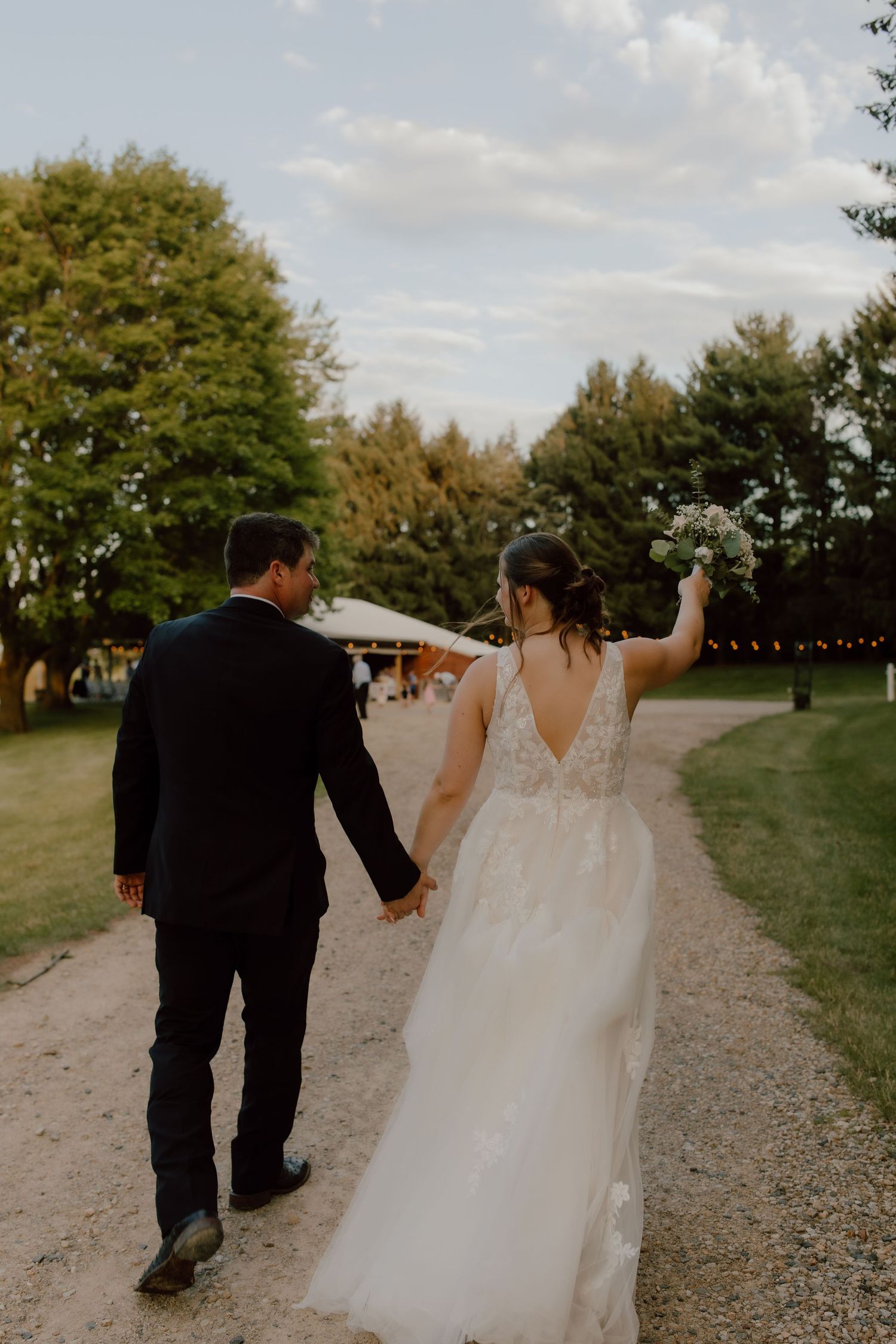 Bride and groom walking hand in hand down a gravel path at an outdoor wedding venue surrounded by lush green trees.