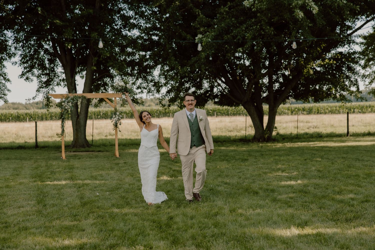 Bride and groom walking hand in hand on green lawn near wooden arch and trees at outdoor wedding.