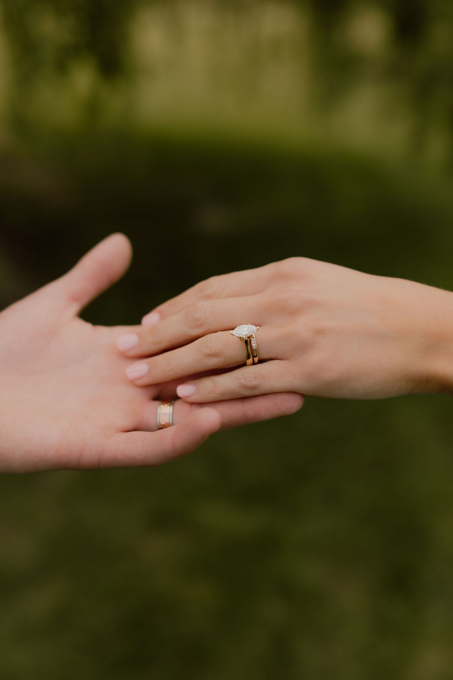 Two hands touching gently, one adorned with a gold engagement ring featuring a white stone, set against a green bokeh background.