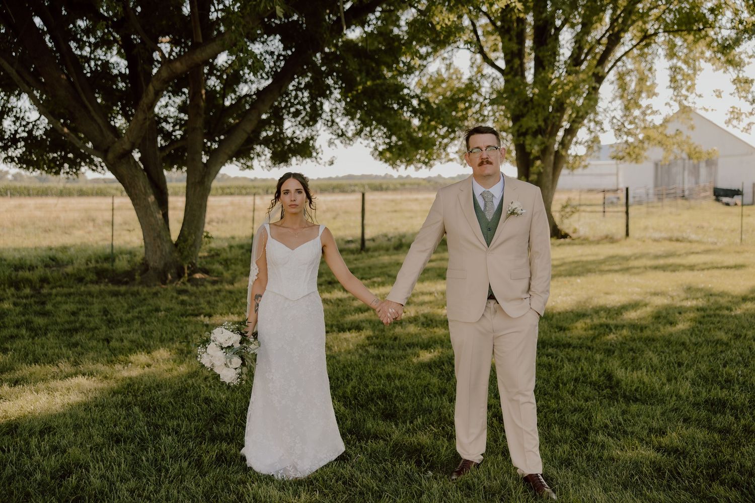 Bride and groom hold hands on green lawn near large trees, she in white gown, he in tan suit.