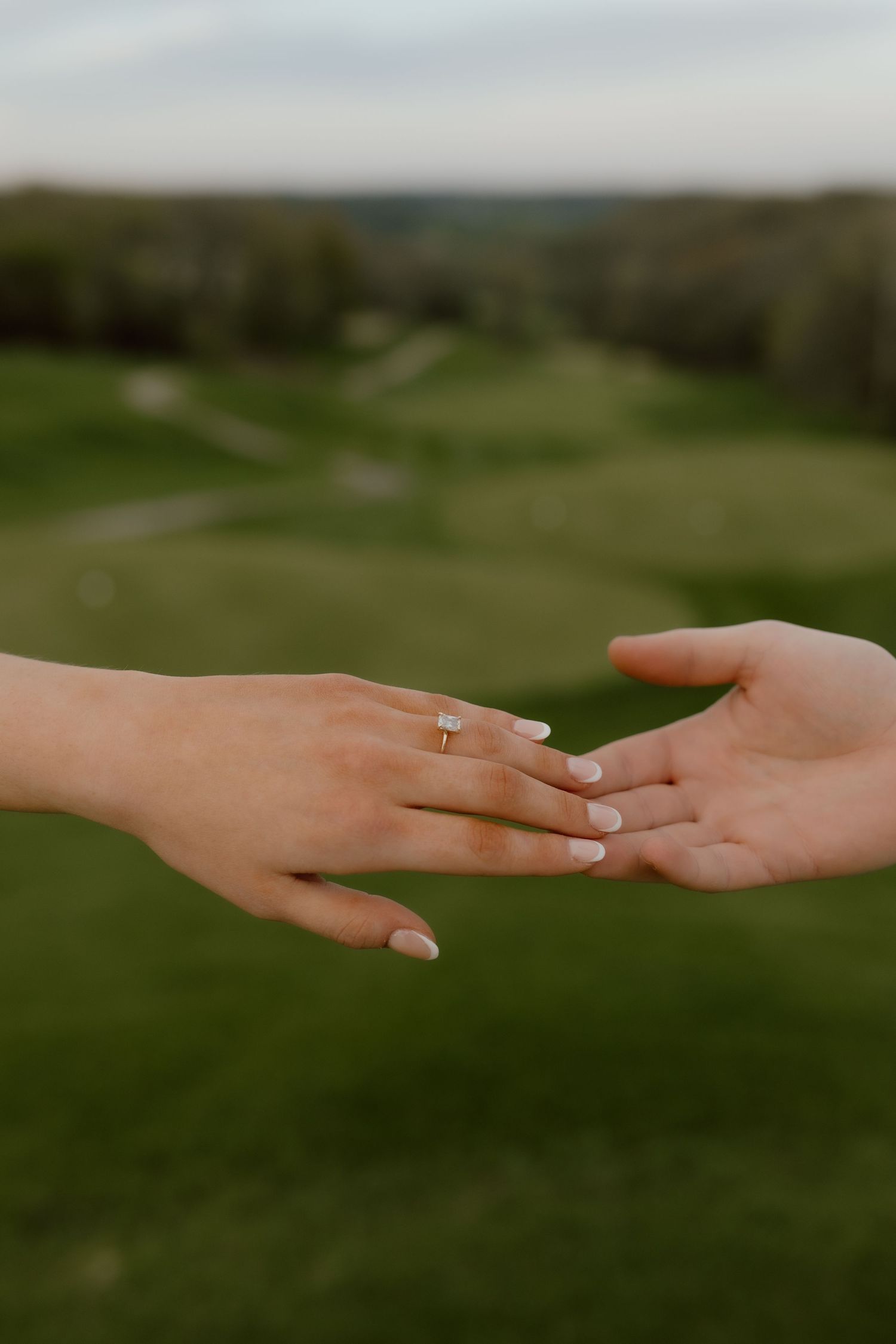 Two hands reaching toward each other with an engagement ring on one finger, set against a lush green golf course backdrop.