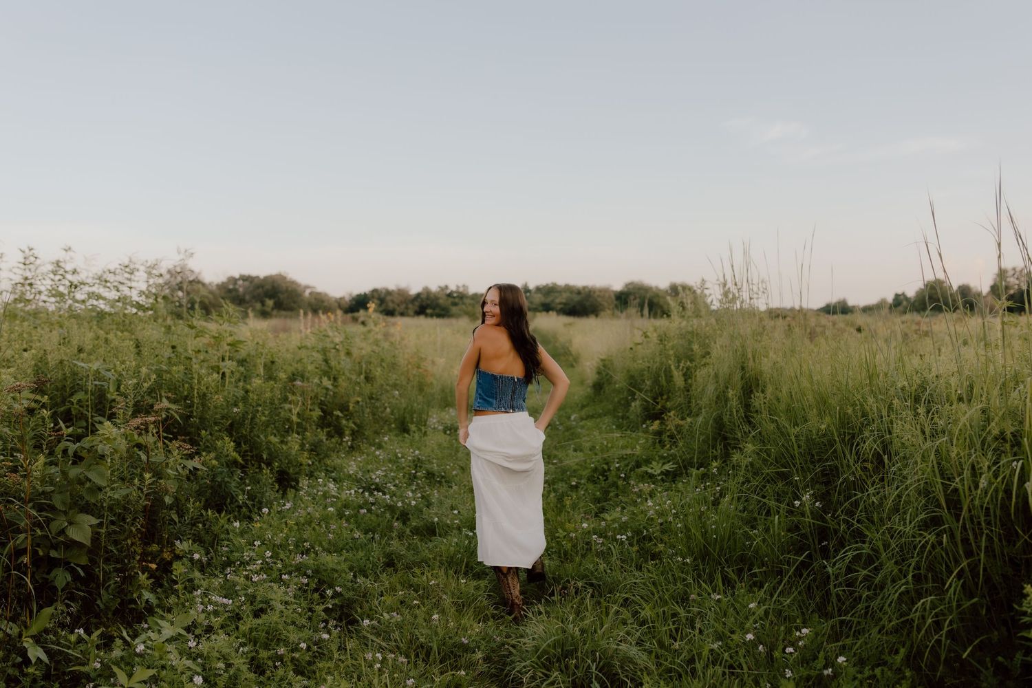 Figure in white skirt standing in tall grass field at sunset.