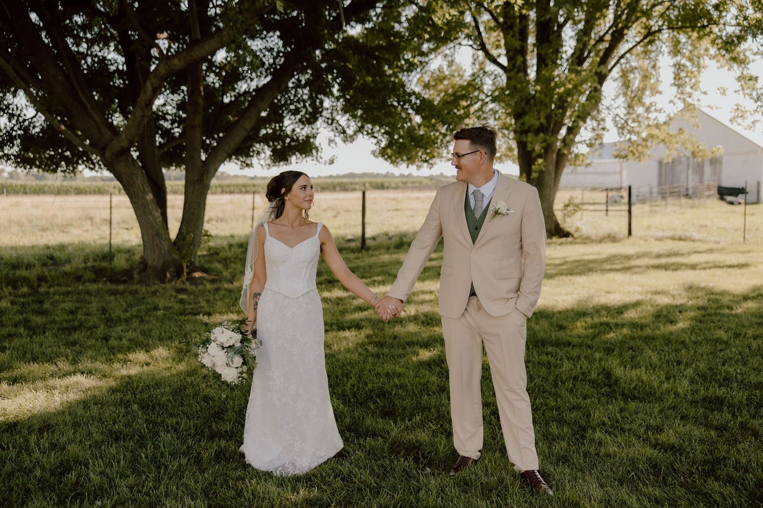 Bride and groom hold hands on green lawn near large tree with white barn in background on wedding day.