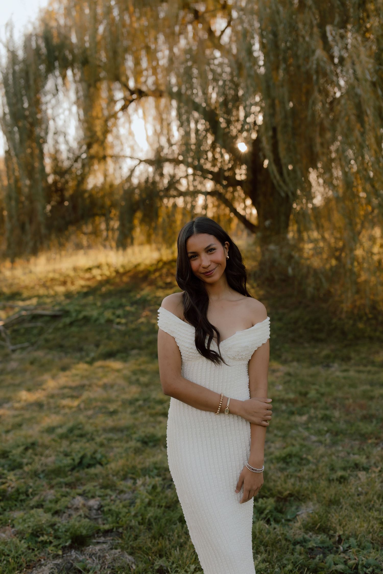 Person in white dress poses gracefully against a backdrop of willow trees at sunset.