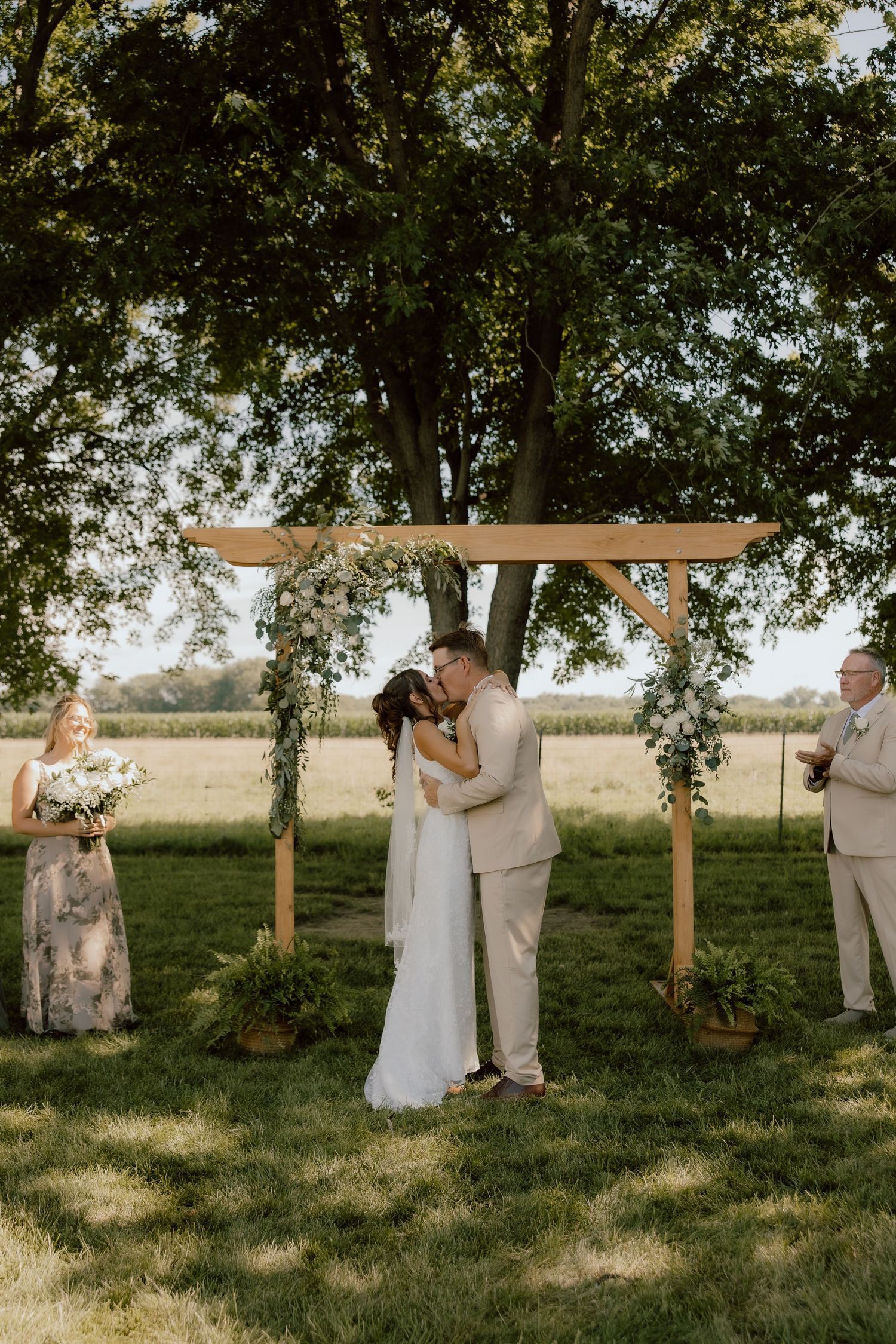 Bride and groom share first kiss under wooden floral arch at outdoor wedding ceremony on lush green lawn.