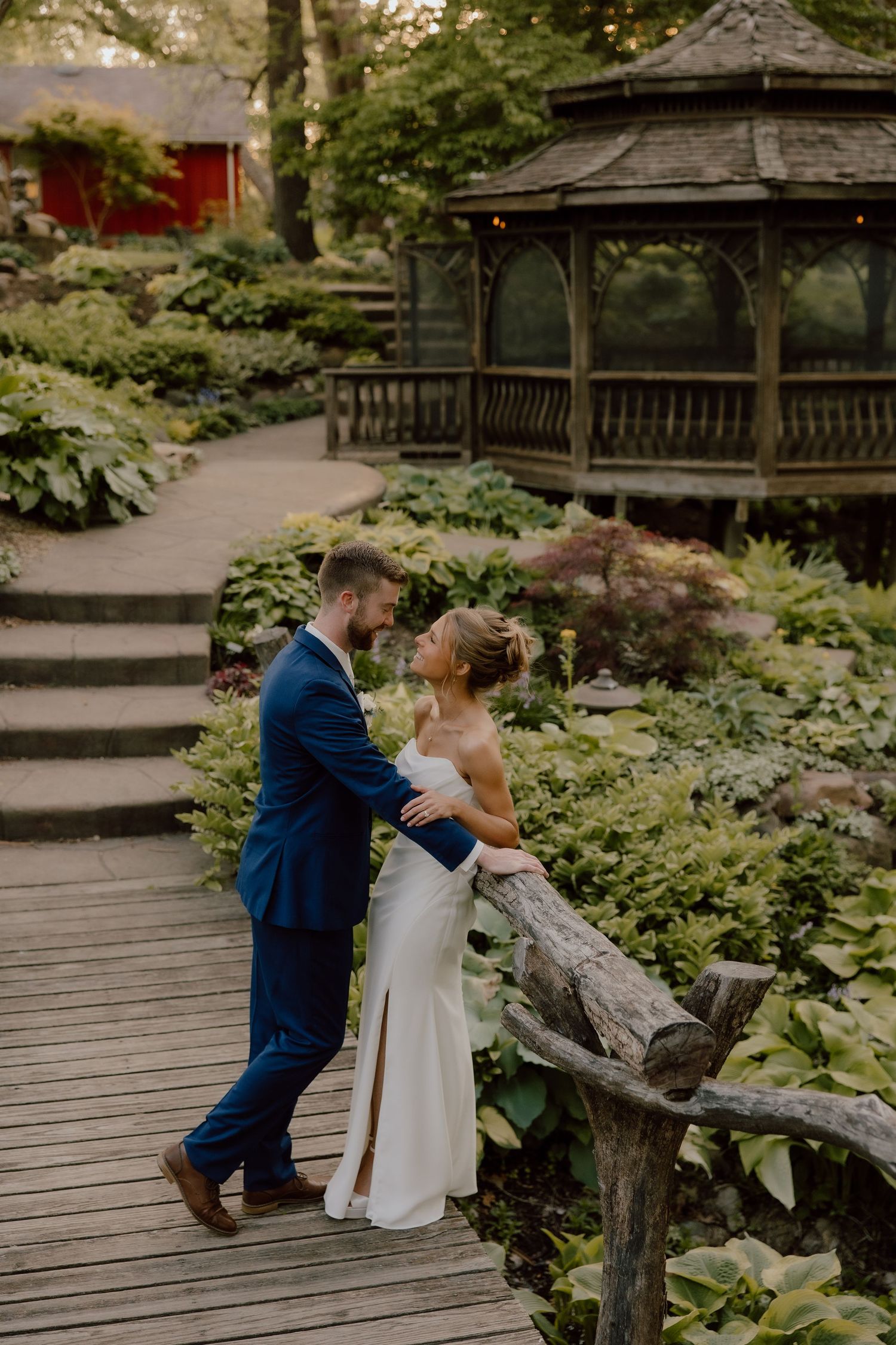 Bride and groom share a romantic moment on a wooden bridge in a lush garden with a gazebo backdrop.