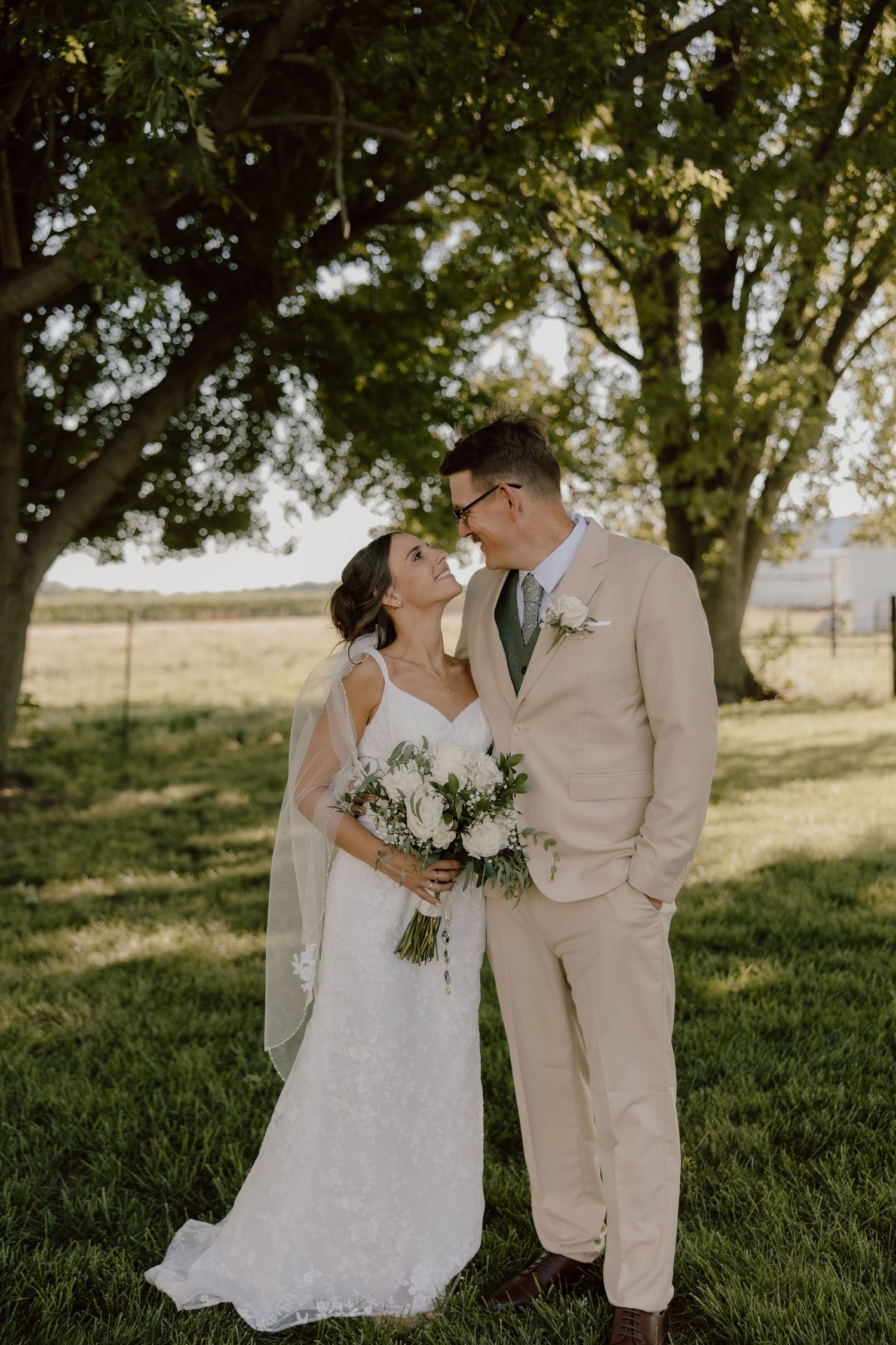 Bride and groom share a romantic moment outdoors, she holds a white bouquet, he wears a tan suit, trees in background.