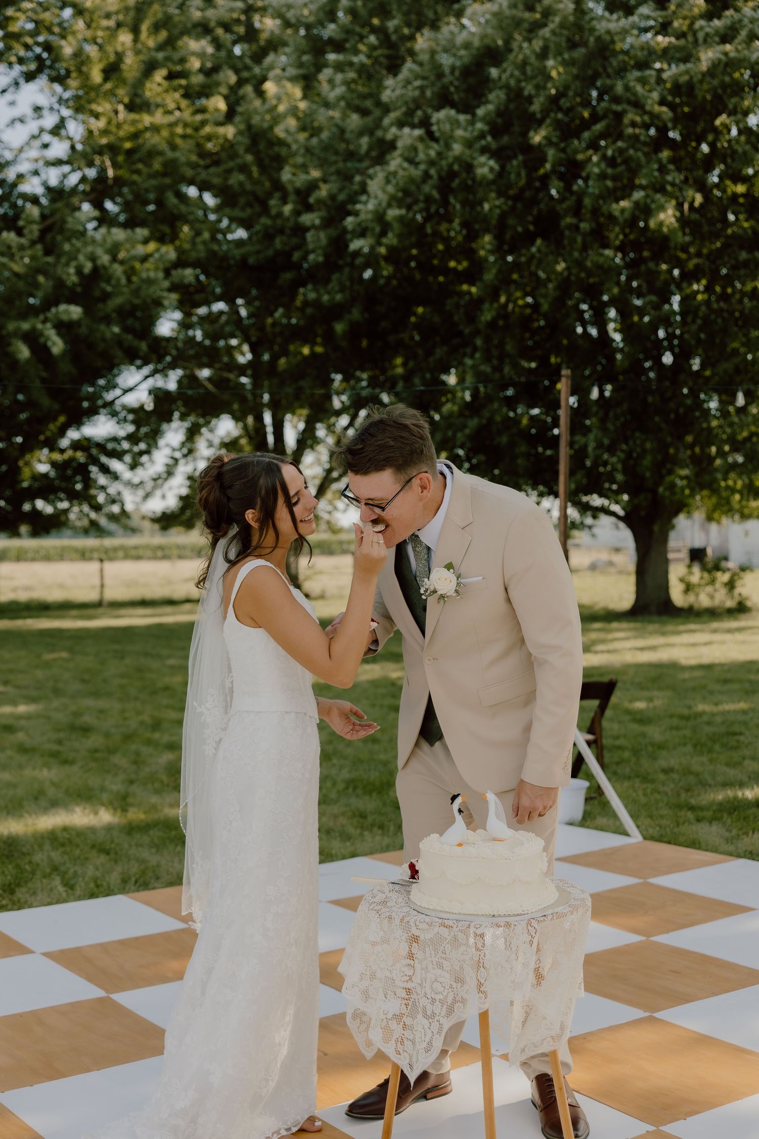 Bride feeds groom wedding cake outdoors on checkered dance floor, surrounded by lush green trees.