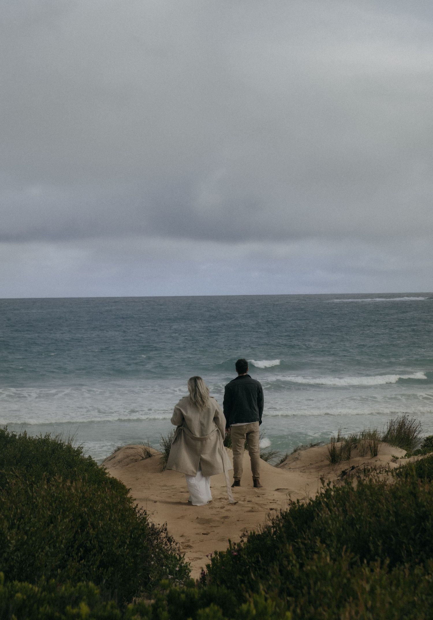 Couple standing together on sandy coastal dunes near Warrnambool overlooking a moody overcast ocean.