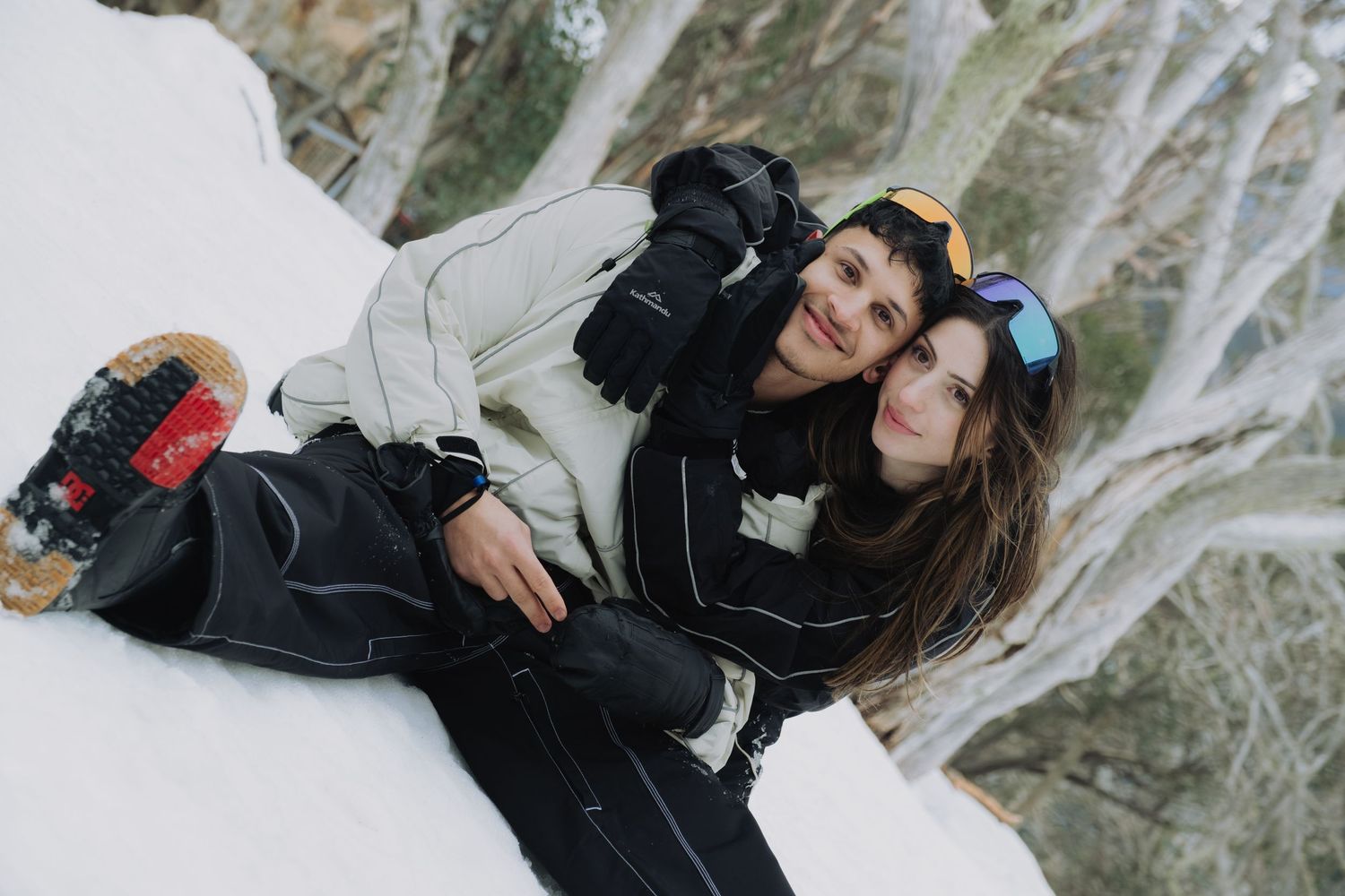 Two snowboarders in winter gear smile together on a snowy slope, wearing goggles and ski jackets.