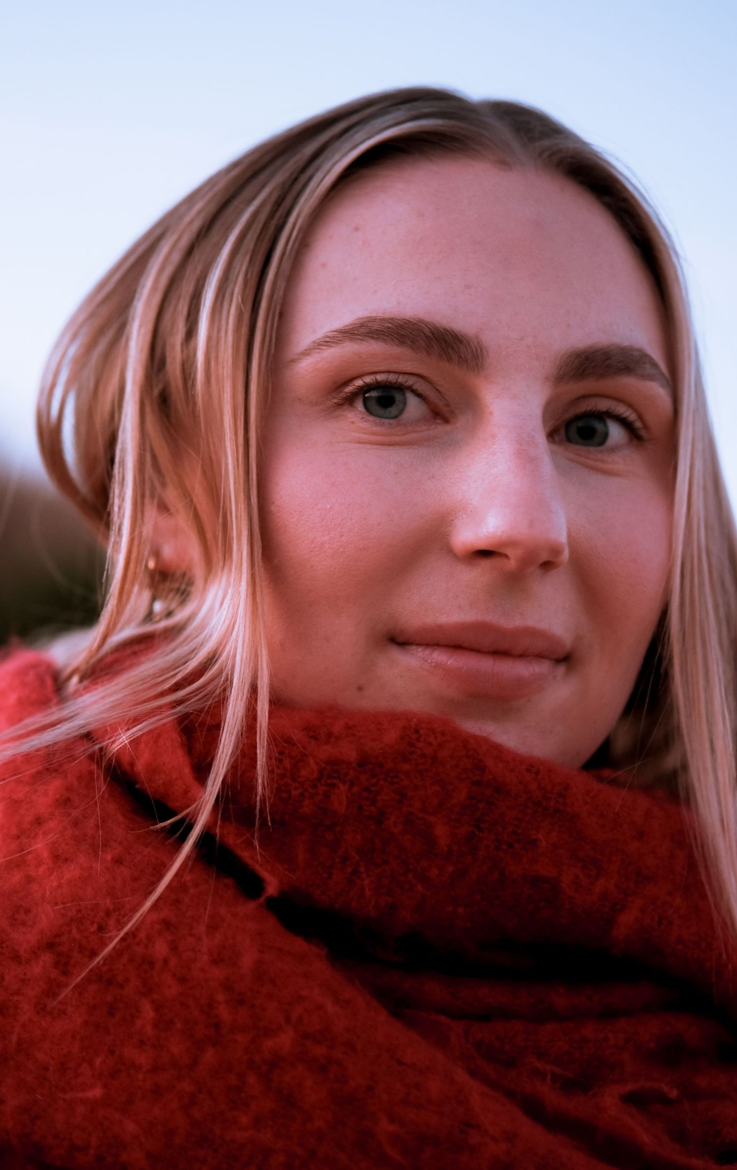 Close-up portrait of a young blonde woman in a red scarf outdoors at dusk.