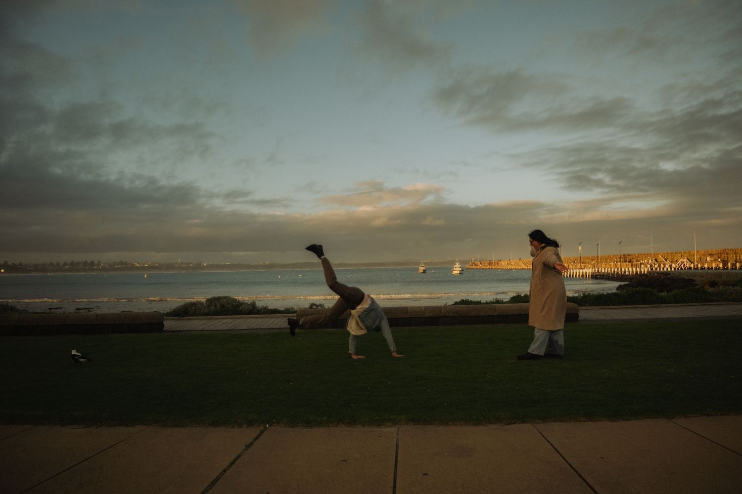 Person doing a cartwheel on grass near waterfront at sunset while another person watches nearby.