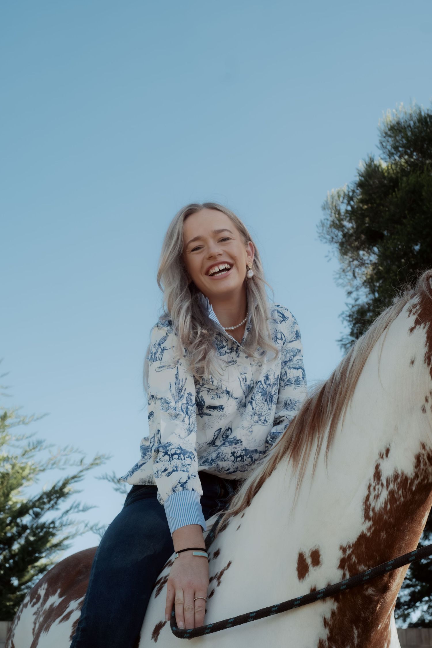 Warrnambool photographer captures smiling blonde woman laughing while riding a horse against a clear blue sky.