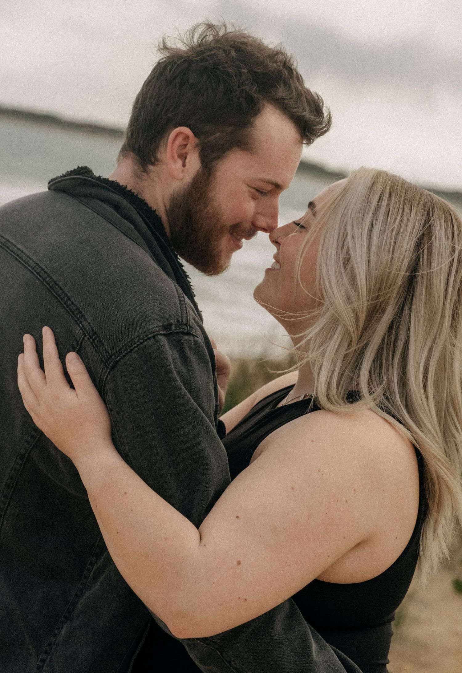Joyful couple almost kissing on a windy beach, woman with blonde hair embracing bearded man.