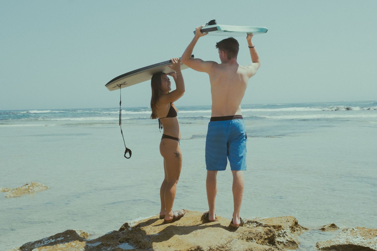 A couple holding surfboards above their heads while standing on rocks overlooking a sunny ocean beach.