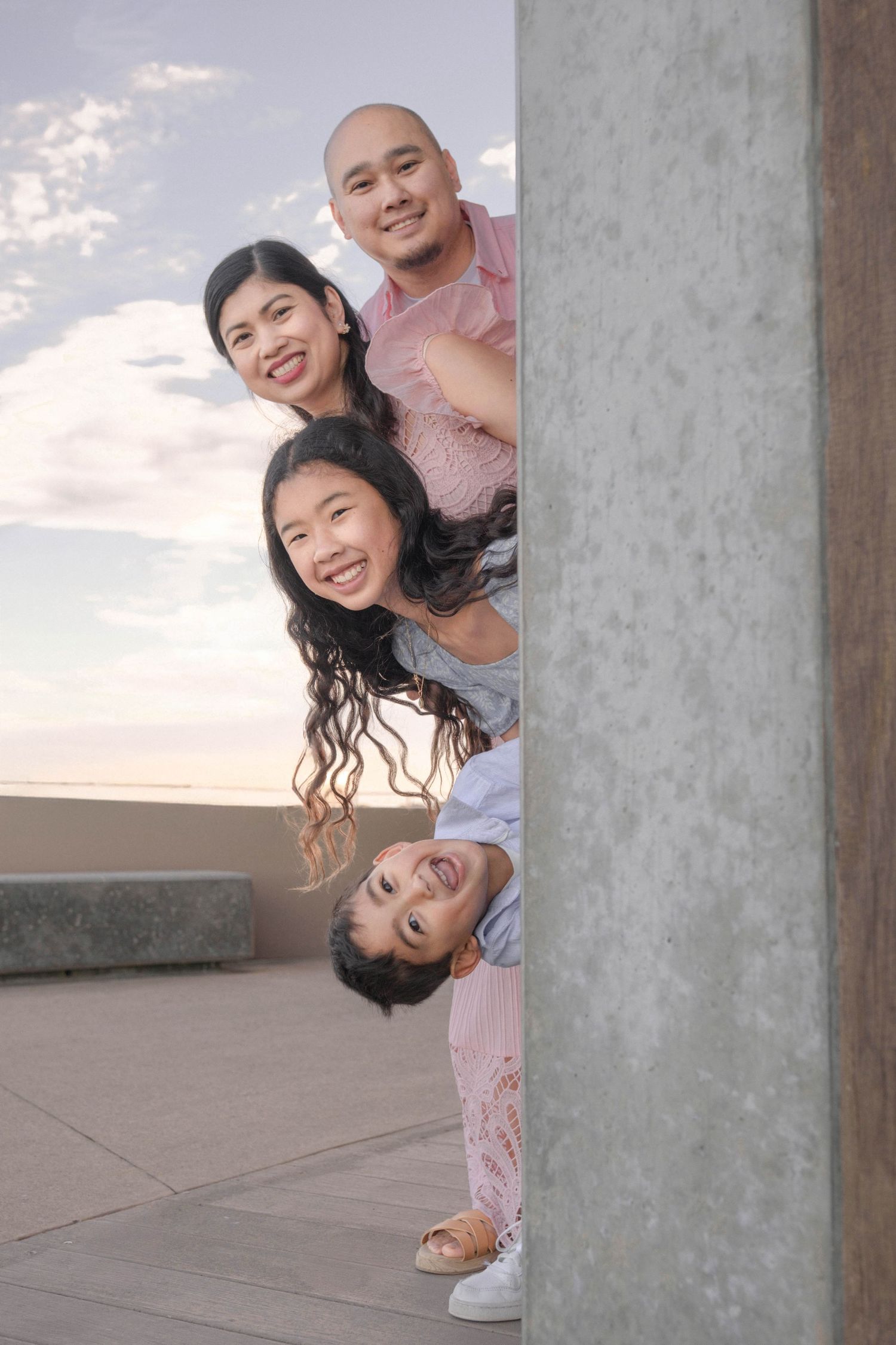A joyful Asian family of four peeking around a concrete pillar outdoors under a cloudy sky.