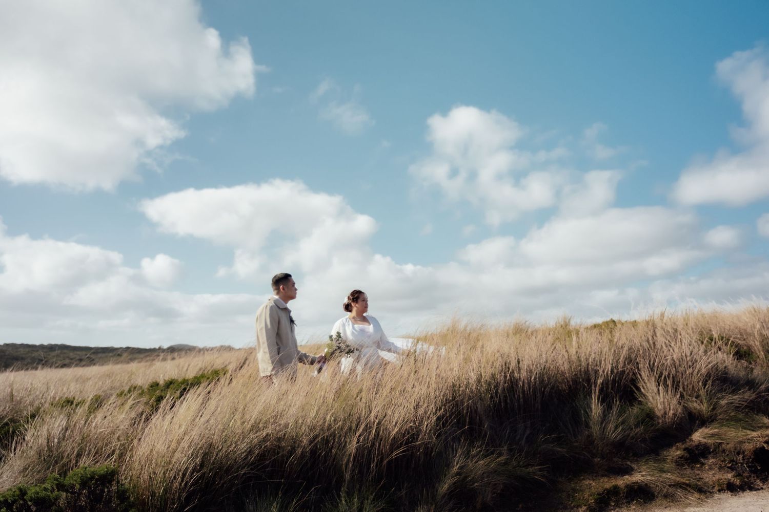 Couple in white wedding attire sitting in tall grass dunes under a blue sky with fluffy clouds.
