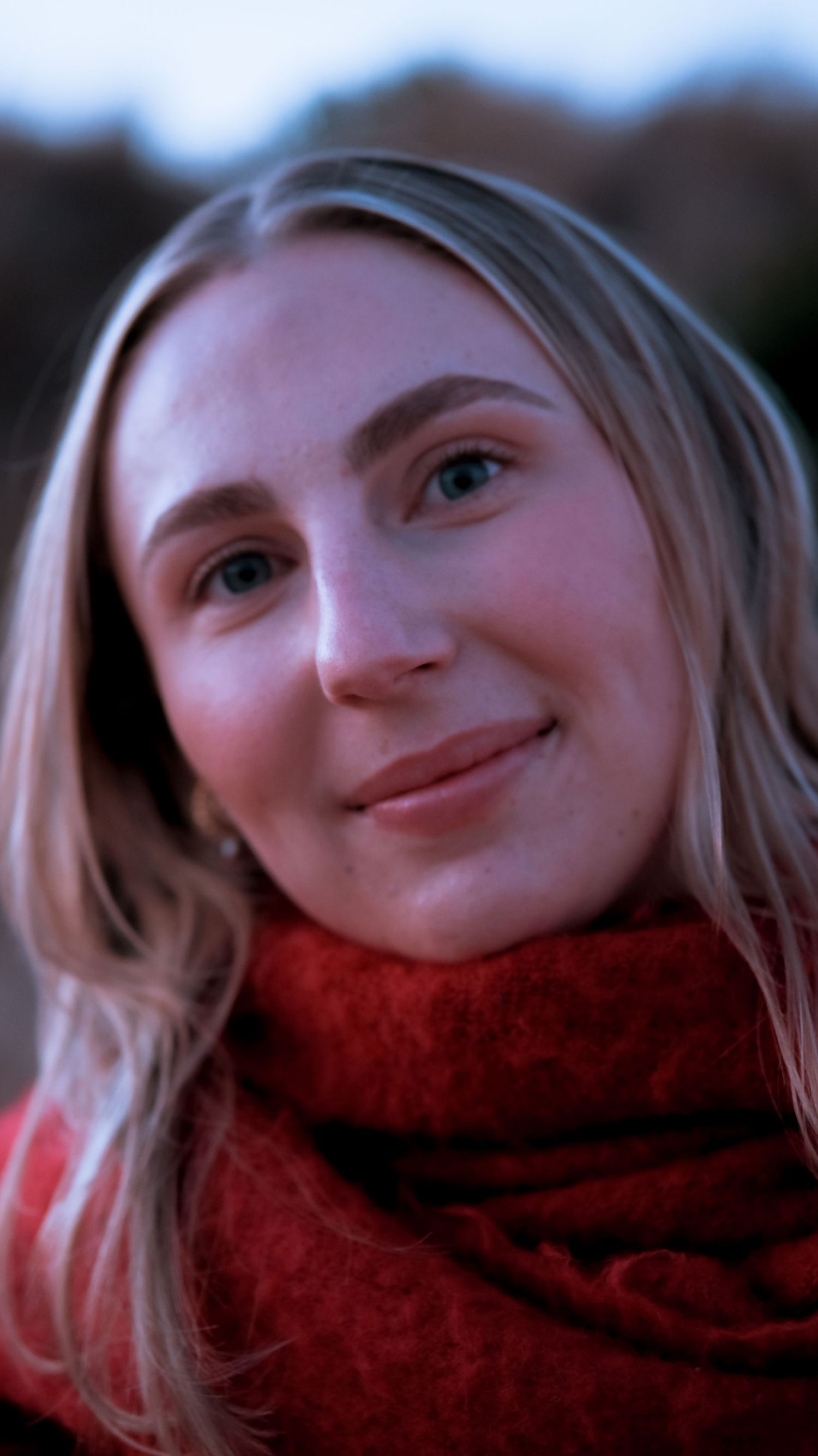 Portrait of a young blonde woman with a subtle smile wearing a red scarf outdoors.