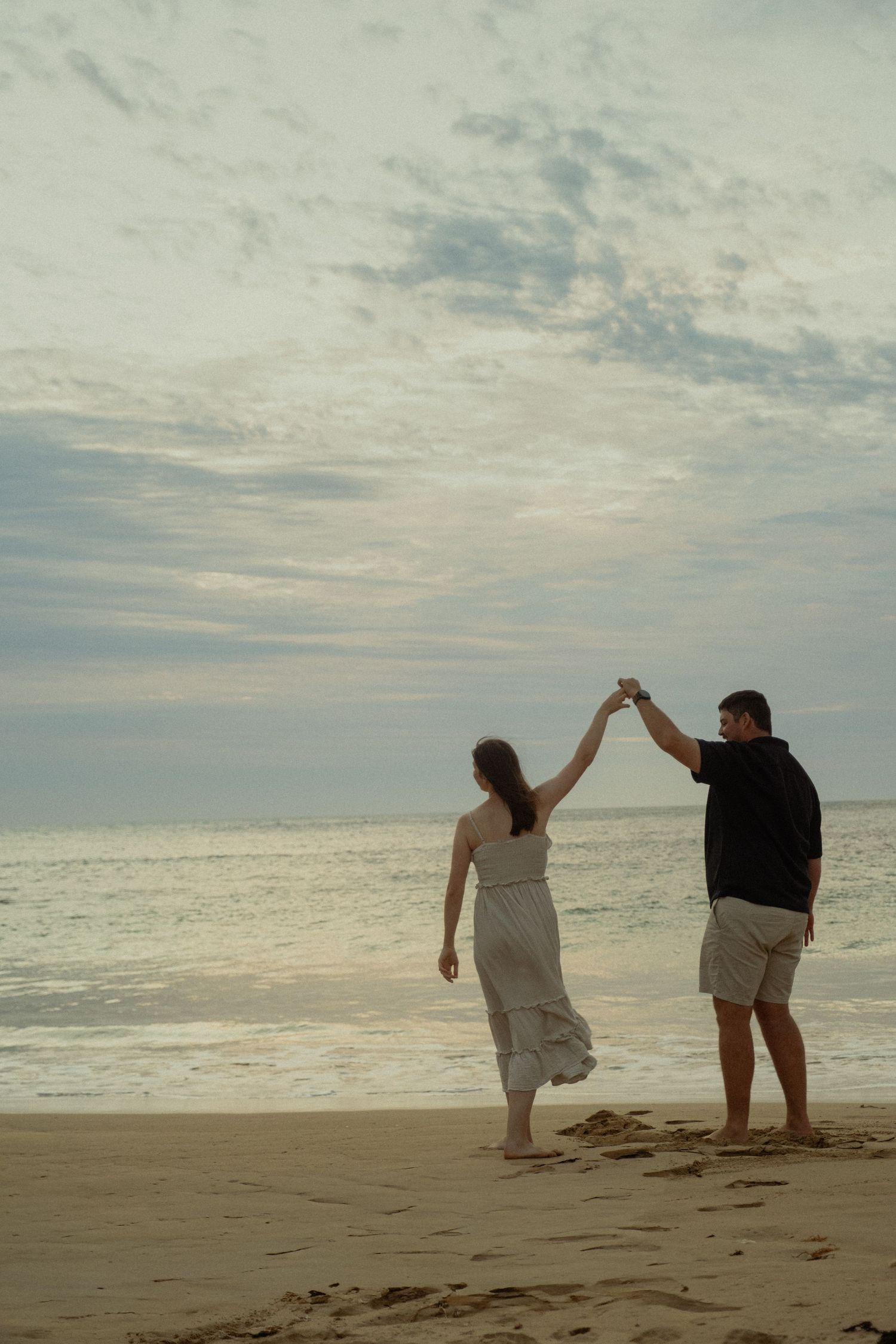 Couple dancing on a beach at sunset, holding hands raised against a cloudy sky.