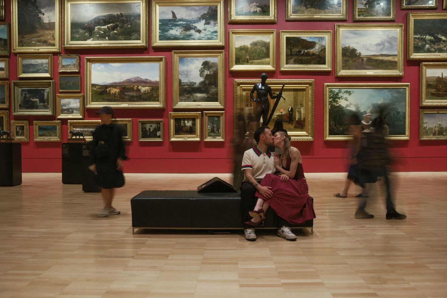 Warrnambool couples photographer captures couple sitting on a bench in a grand art gallery with red walls covered in framed paintings.