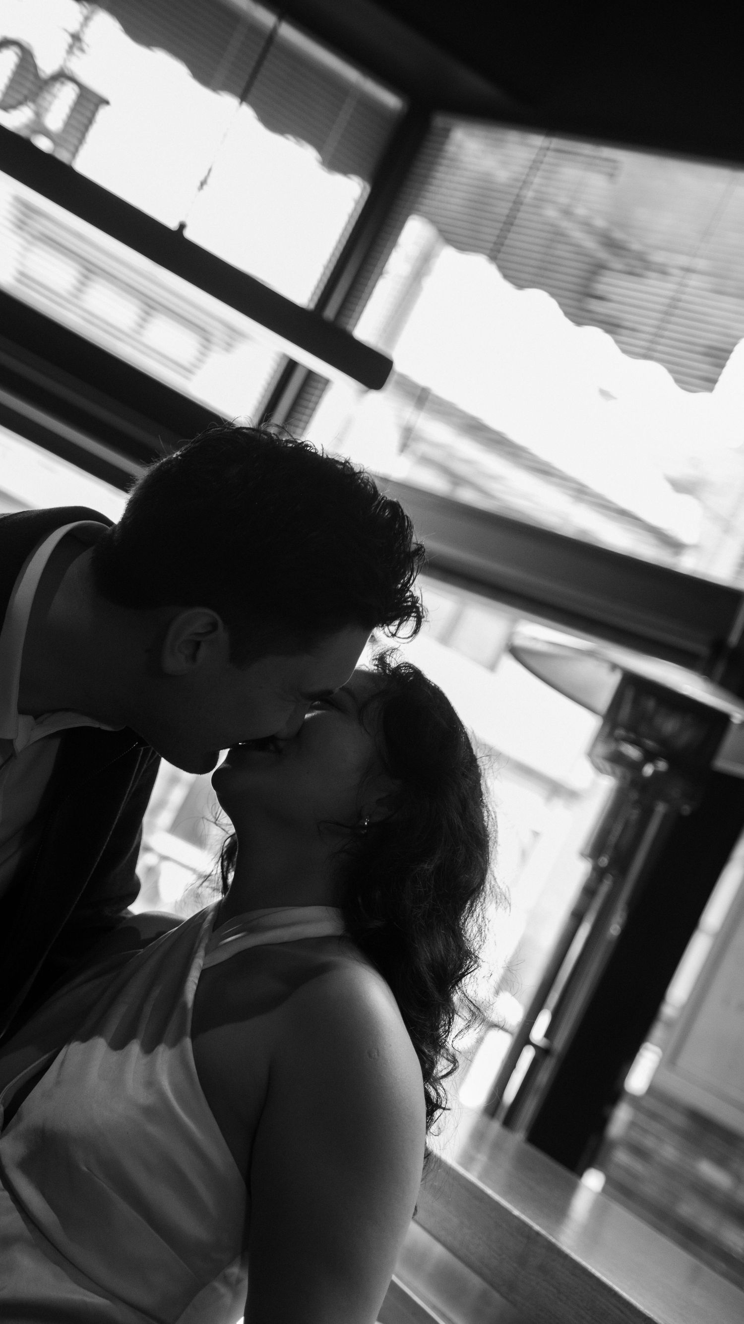 Black and white photo of a couple sharing a romantic kiss on a city street, captured in soft natural light.
