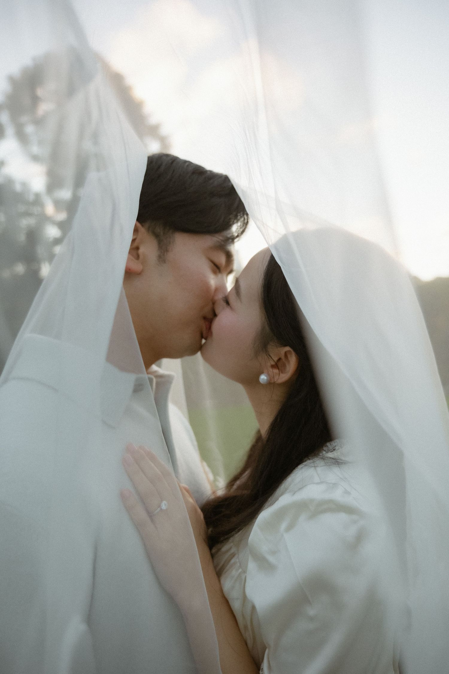 A couple shares a romantic kiss under a flowing white veil, the bride wearing a pearl earring and engagement ring.