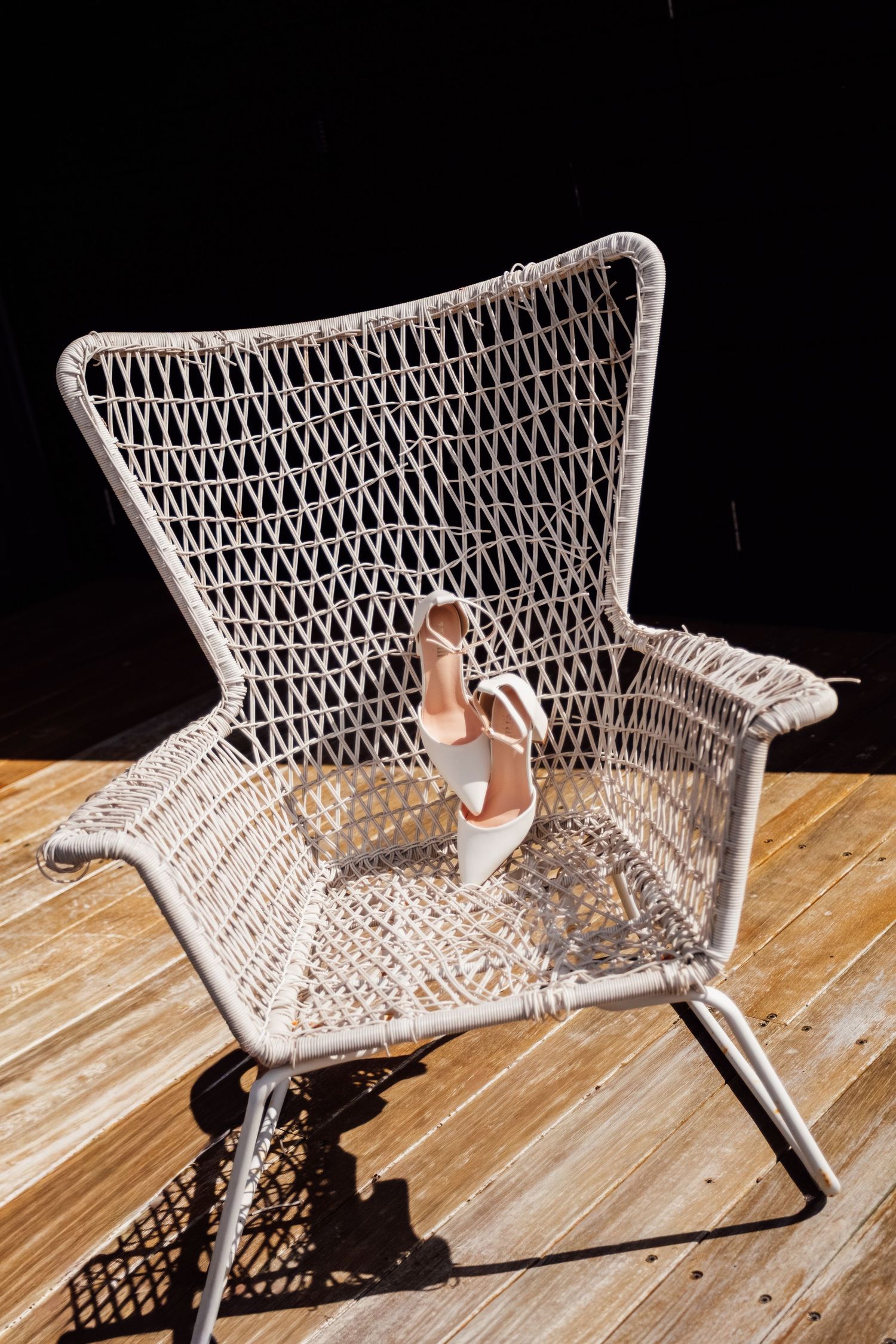 White wicker chair on wooden deck with elegant bridal heels placed on seat against dark background.
