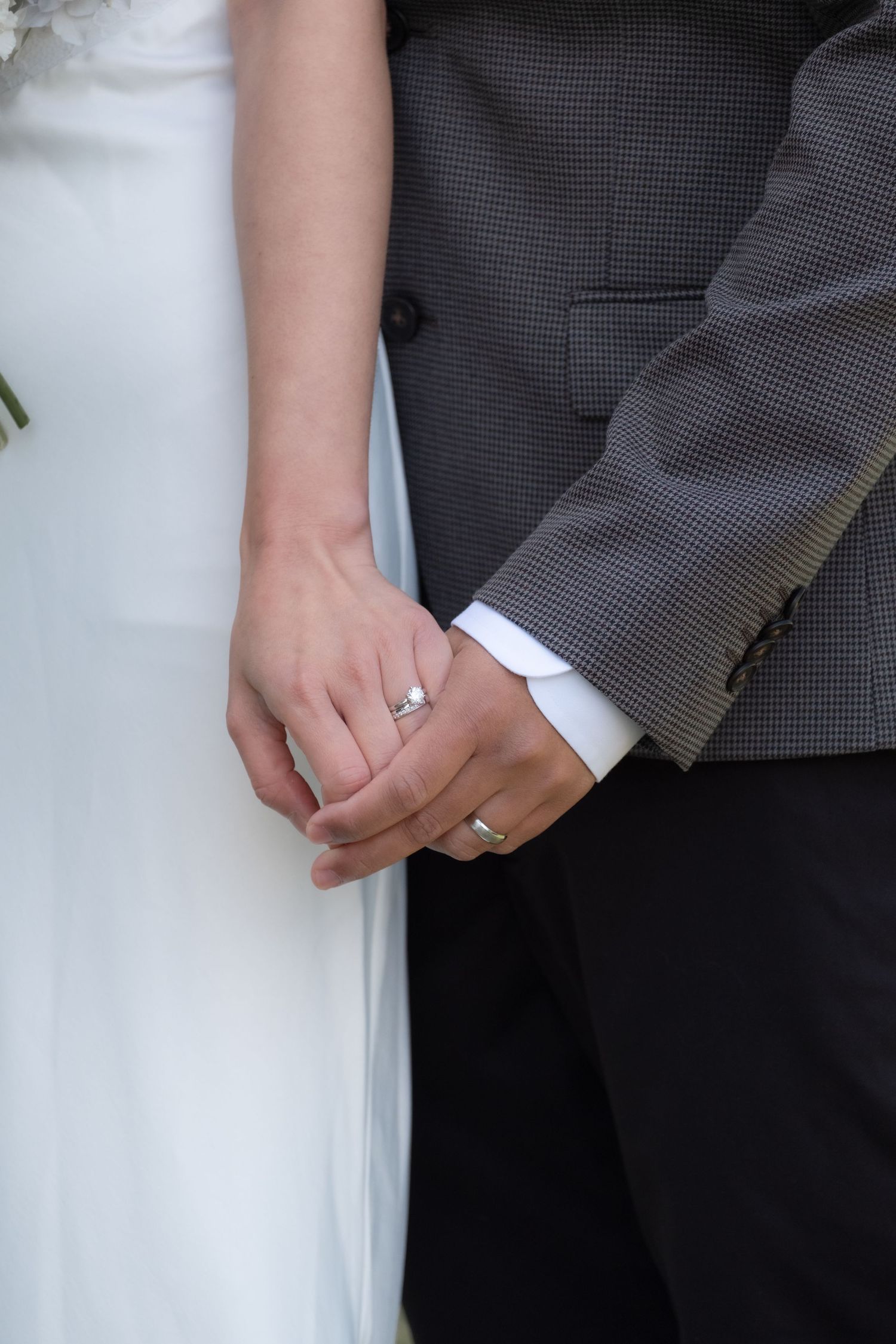 Newlywed couple holding hands showing wedding rings, bride in white gown and groom in gray suit.