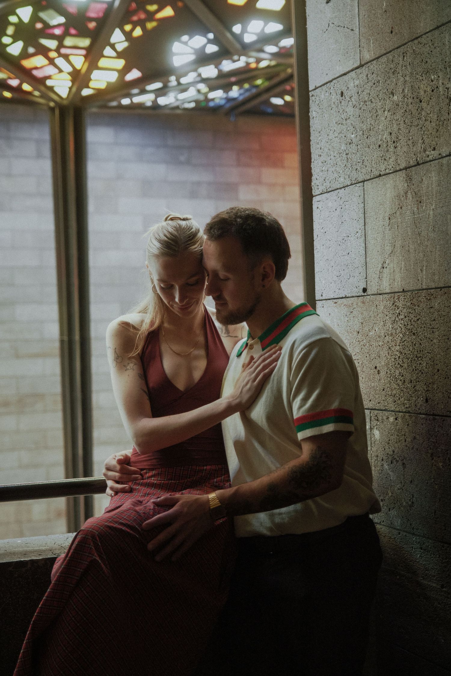 Warrnambool couples photographer captures couple sitting intimately together beneath colorful stained glass window indoors.