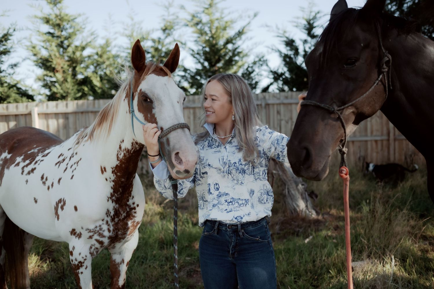 Warrnambool photographer captures cheerful blonde woman standing between two horses at a sunny outdoor ranch.