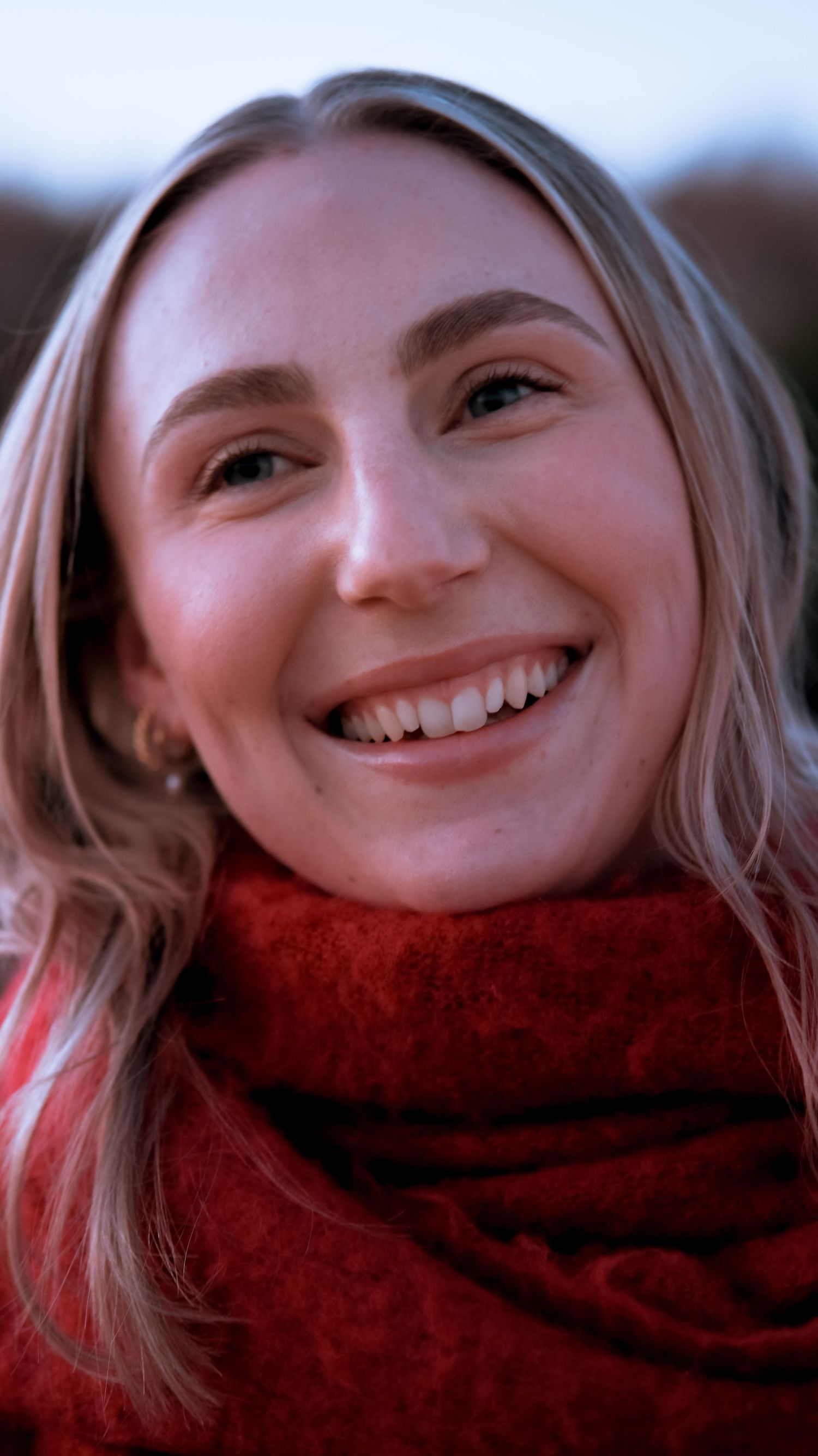 Smiling young blonde woman wearing a cozy red scarf photographed outdoors in natural light.