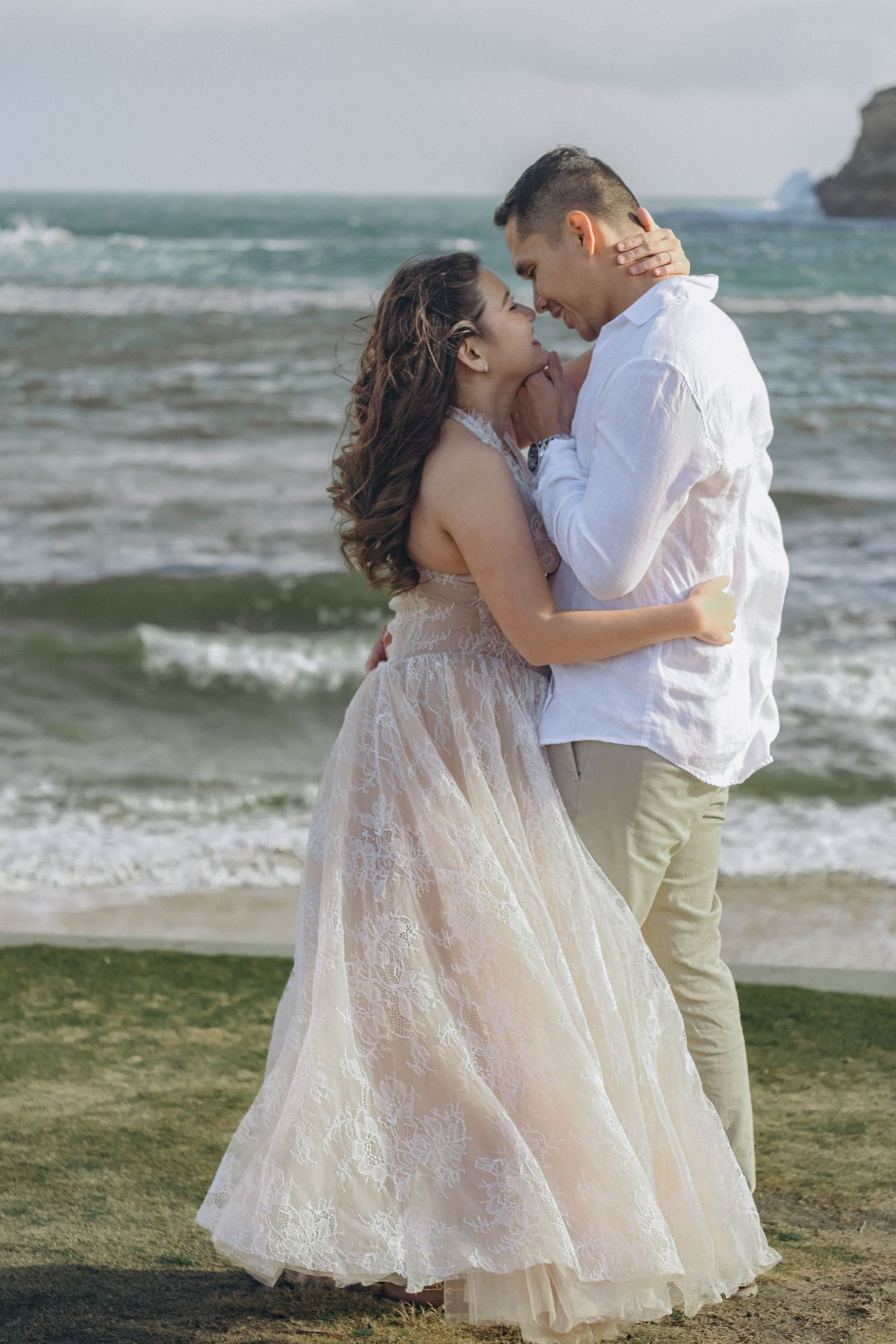 Couple embracing on a rocky beach, woman in lace gown, man in white shirt, ocean waves in background.