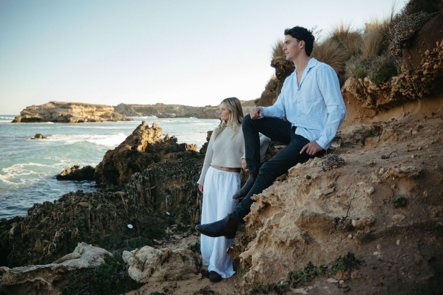 Couple sitting on rocky coastal cliffs at sunset, woman in white dress, man in white shirt, ocean waves in background.