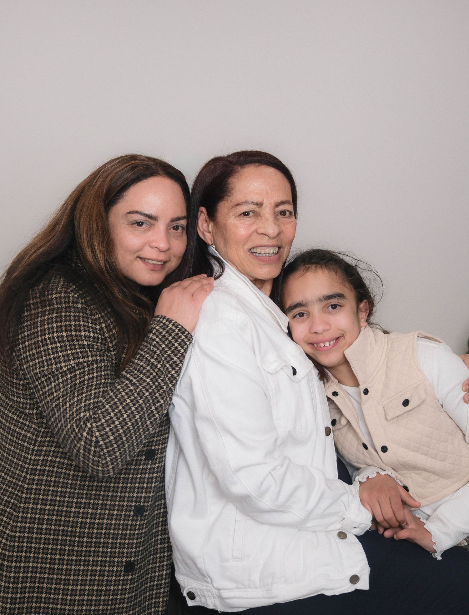 Three generations of women smiling together in casual outfits against gray background.