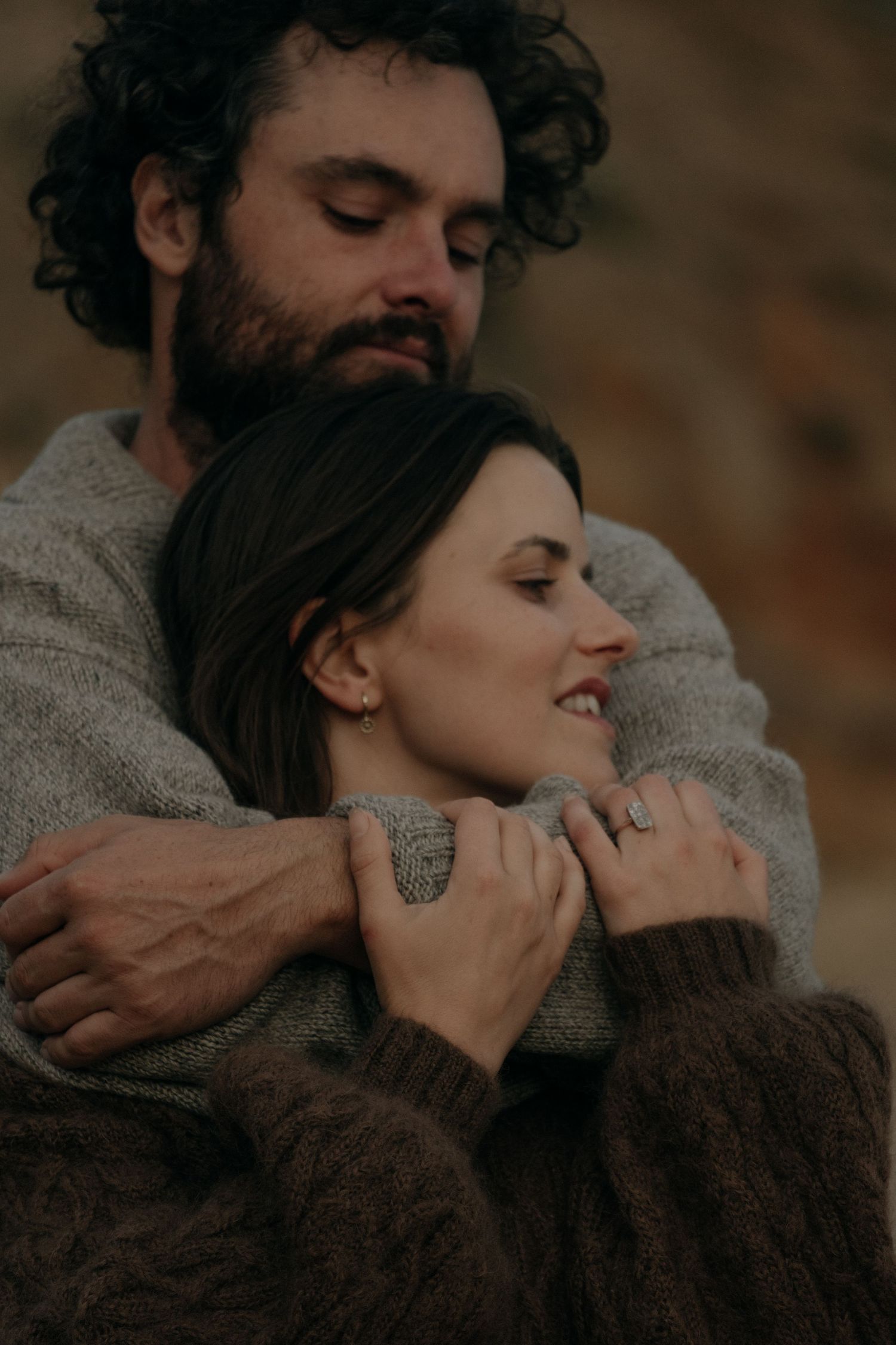 Romantic couple embracing outdoors, man in brown sweater wrapping arms around smiling woman.