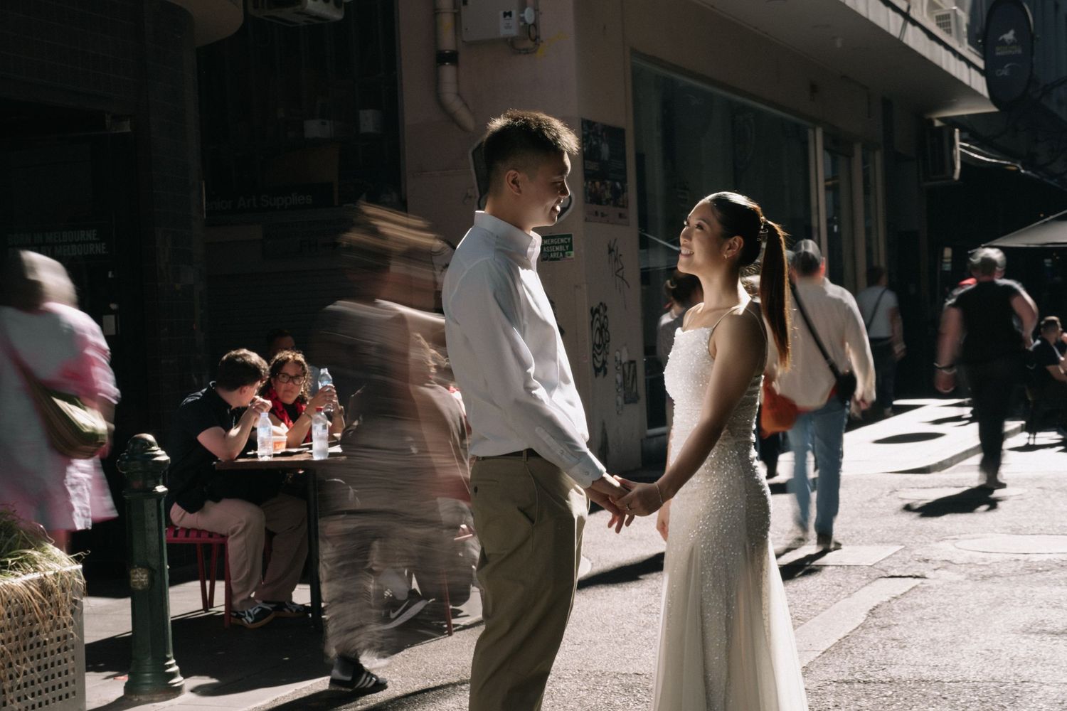 Couple in wedding attire holding hands on a sunny urban street, with blurred pedestrians and a café in the background.