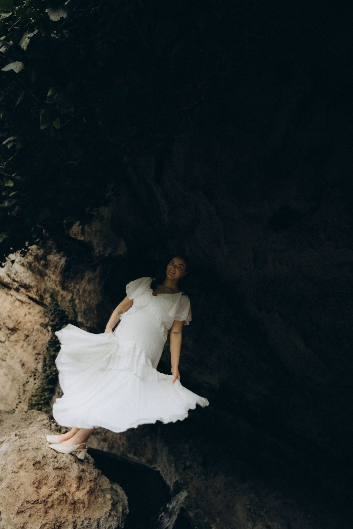 Woman in flowing white dress standing on rocky terrain in a dark cave setting.