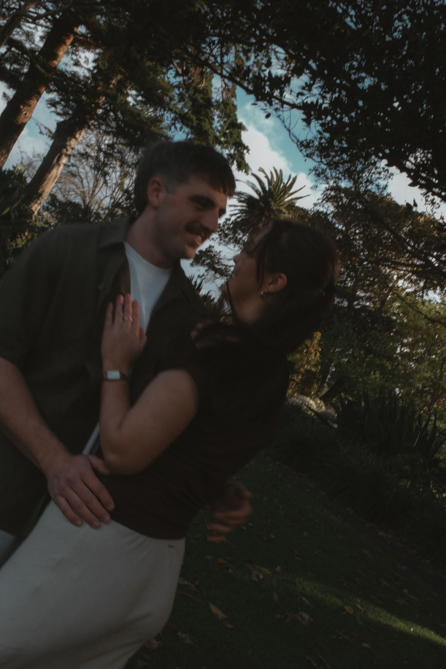 Smiling couple embracing and gazing at each other in a lush garden surrounded by tall green trees.